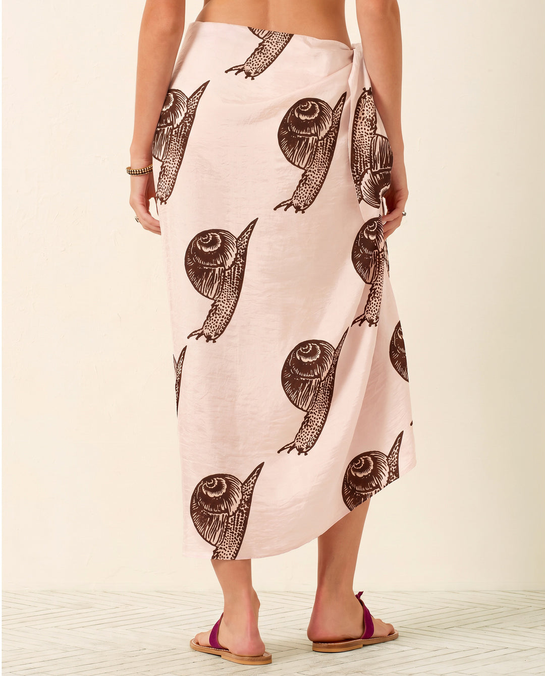 A person wearing the Soraya Snail Study Silk Sarong—a light pink, hand-block-printed piece featuring a brown snail pattern—stands barefoot on a light-colored floor, facing away from the camera.