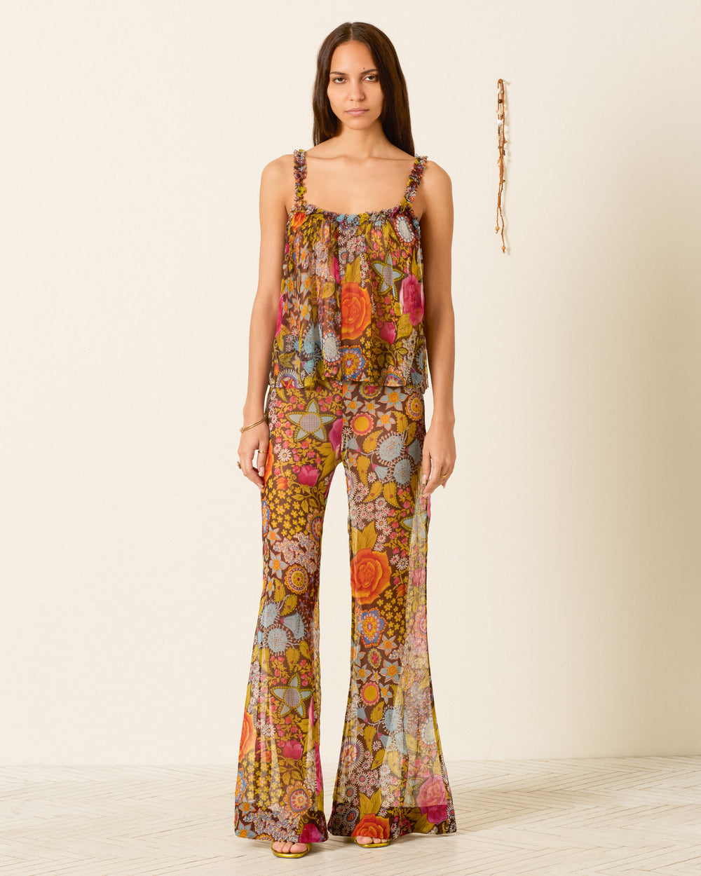 A woman stands against a plain wall wearing the Tati Bloom Mirage Top—a sleeveless, floral camisole with ruffled trim in bright colors crafted from airy viscose-chiffon—paired with matching flared pants.