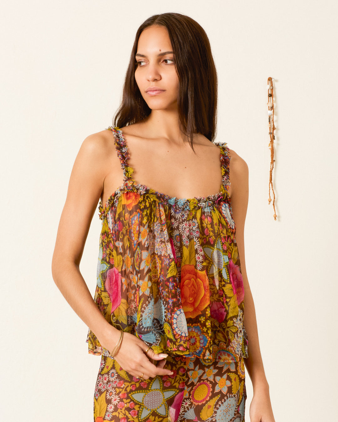 A woman stands against a plain light-colored wall, wearing the Tati Bloom Mirage Top—a floral-patterned, sleeveless, sheer viscose-chiffon piece with delicate ruffled trim—paired with matching pants; a hanging accessory is visible in the background.