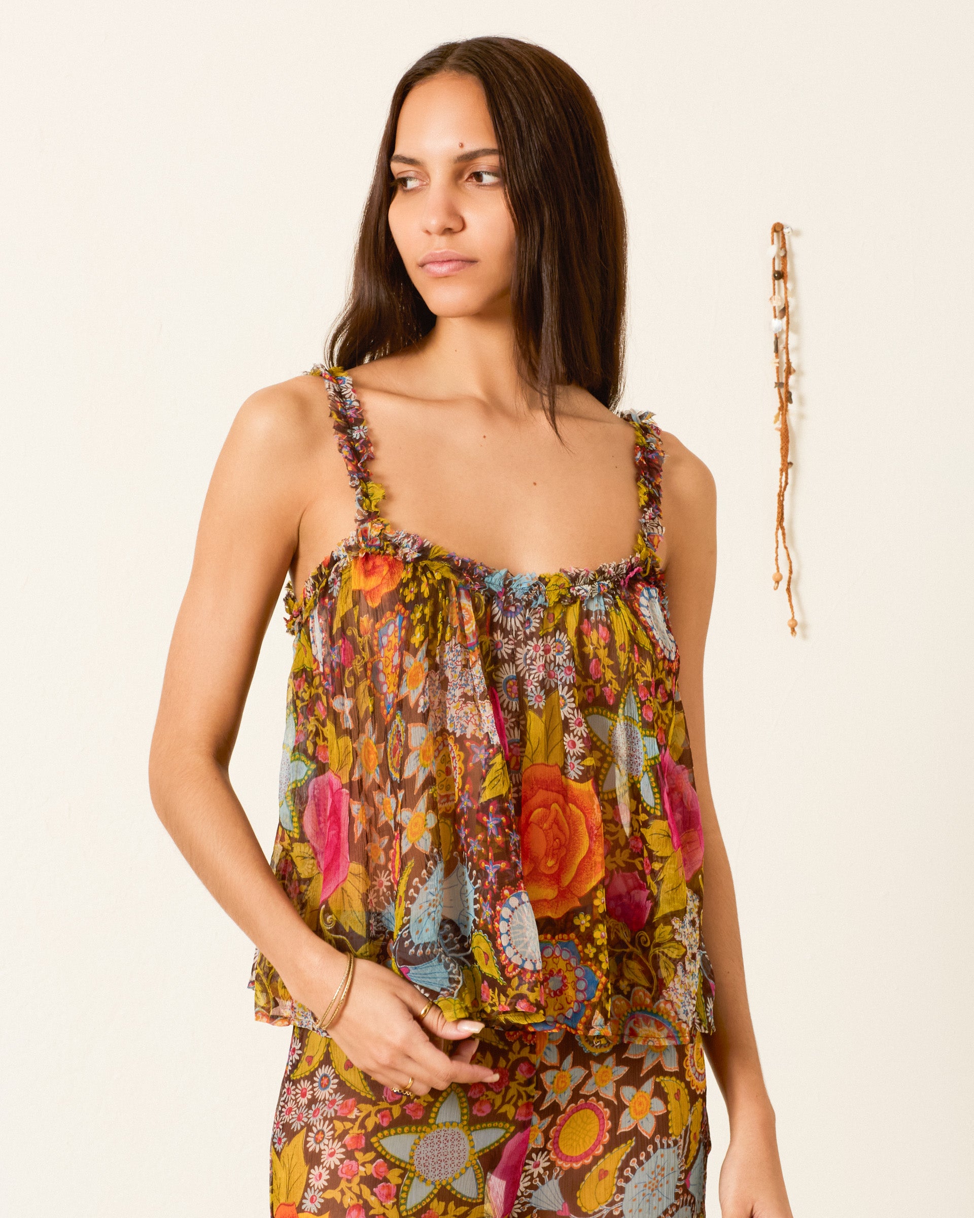 A woman stands against a plain light-colored wall, wearing the Tati Bloom Mirage Top—a floral-patterned, sleeveless, sheer viscose-chiffon piece with delicate ruffled trim—paired with matching pants; a hanging accessory is visible in the background.