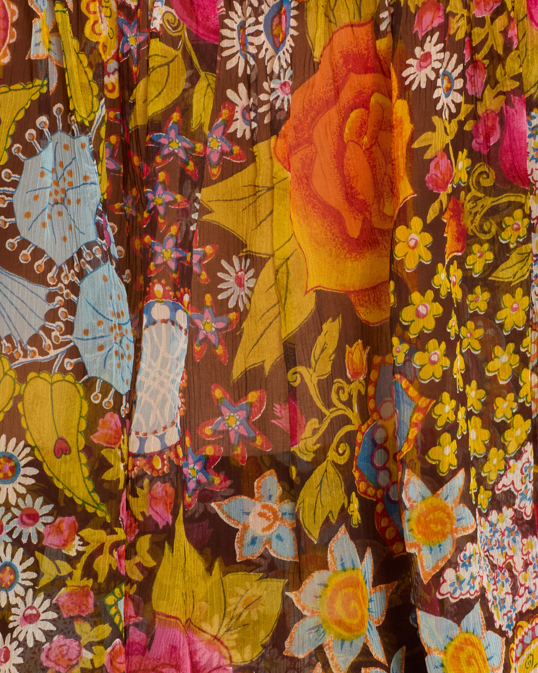 The Tati Bloom Mirage Top is crafted from a translucent viscose-chiffon fabric adorned with a vibrant floral pattern—showcasing large orange and yellow roses, blue accents, and delicate pink, yellow, and white blossoms on a brown background—making it perfect for creating a delicate camisole with ruffled trim.