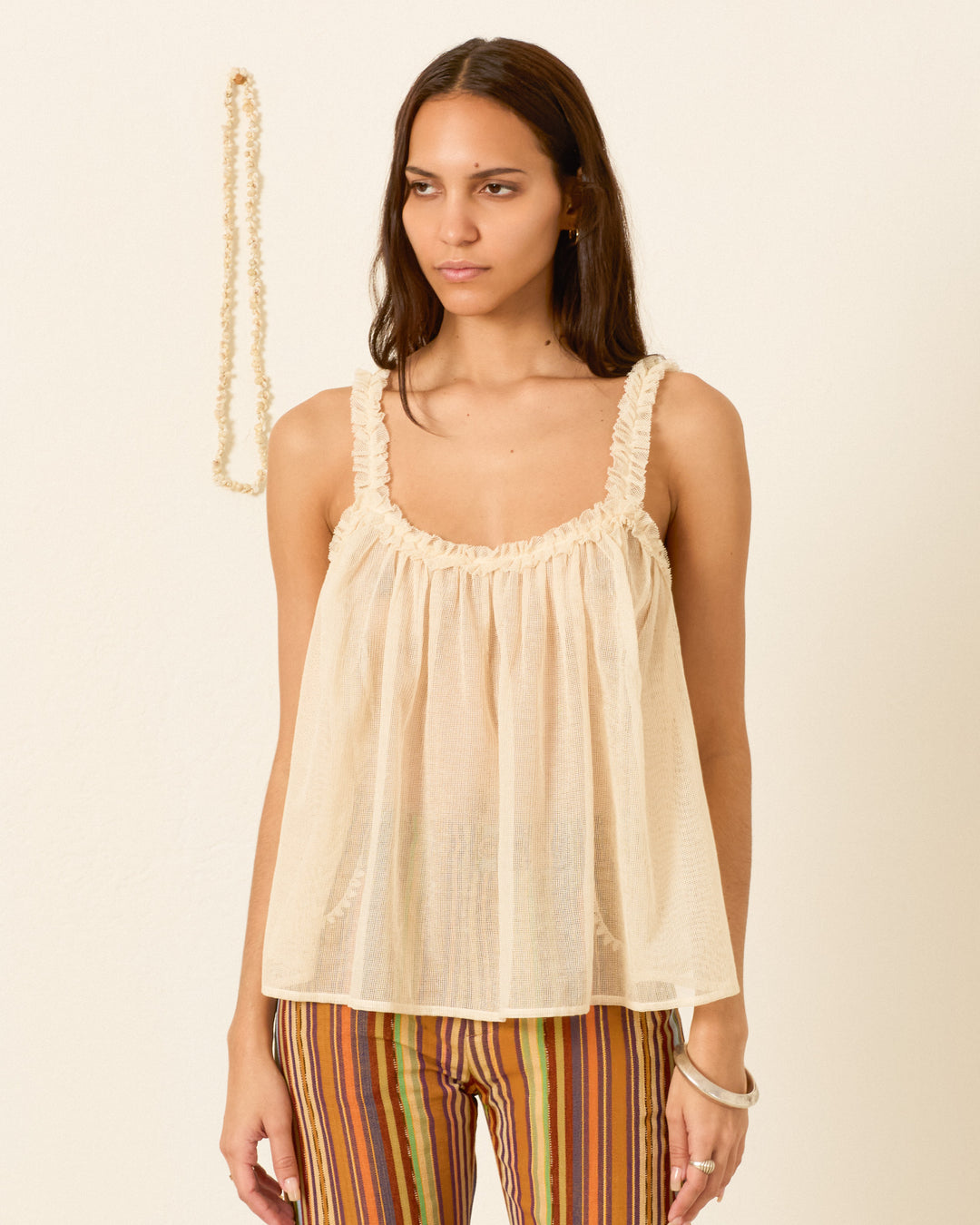 A woman stands against a plain wall wearing the Tati Pearl Weave Top, a loose, sleeveless, cream-colored cotton mesh style, paired with multicolored striped pants. A necklace hangs on the wall behind her.