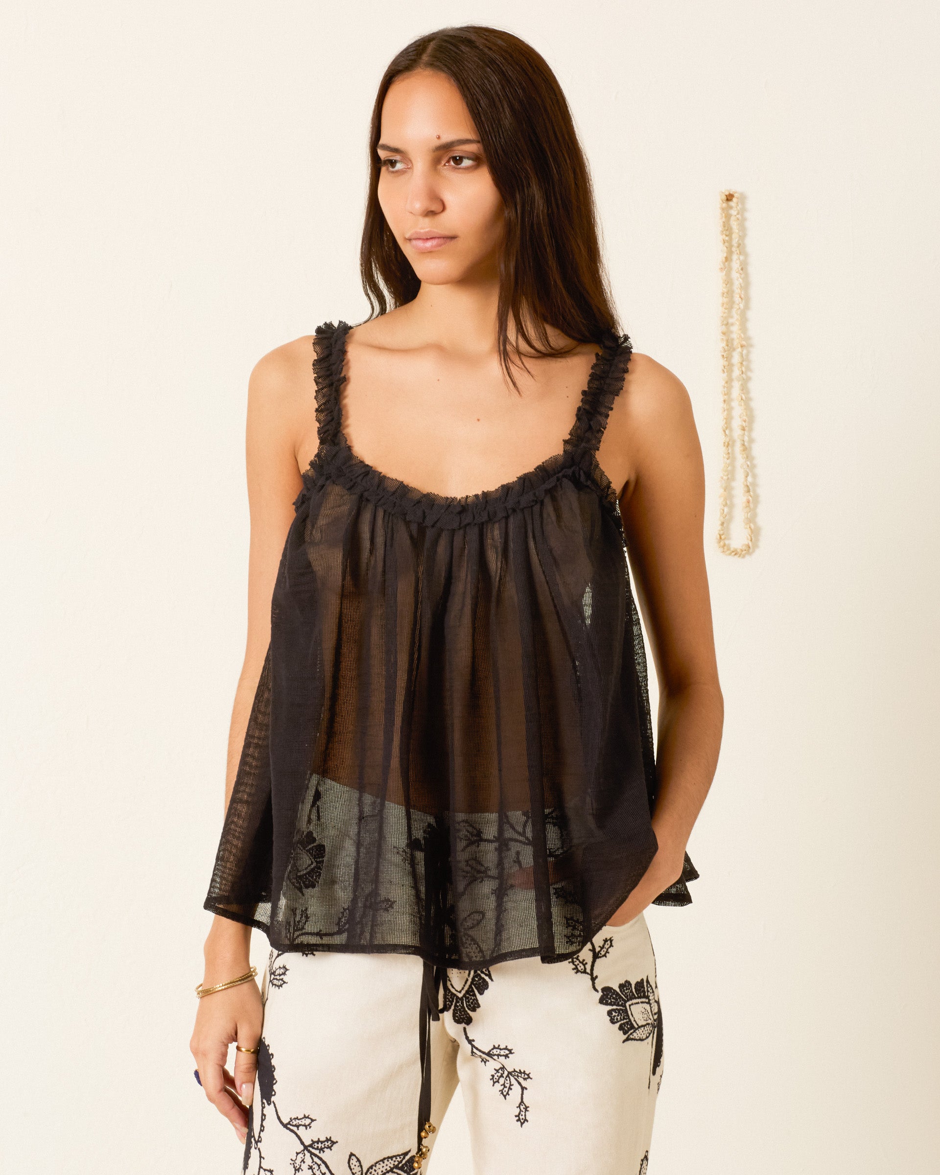 A woman wears the Tati Onyx Weave Top, a sheer camisole with ruffled trim straps, paired with white pants featuring black floral embroidery against a plain background.