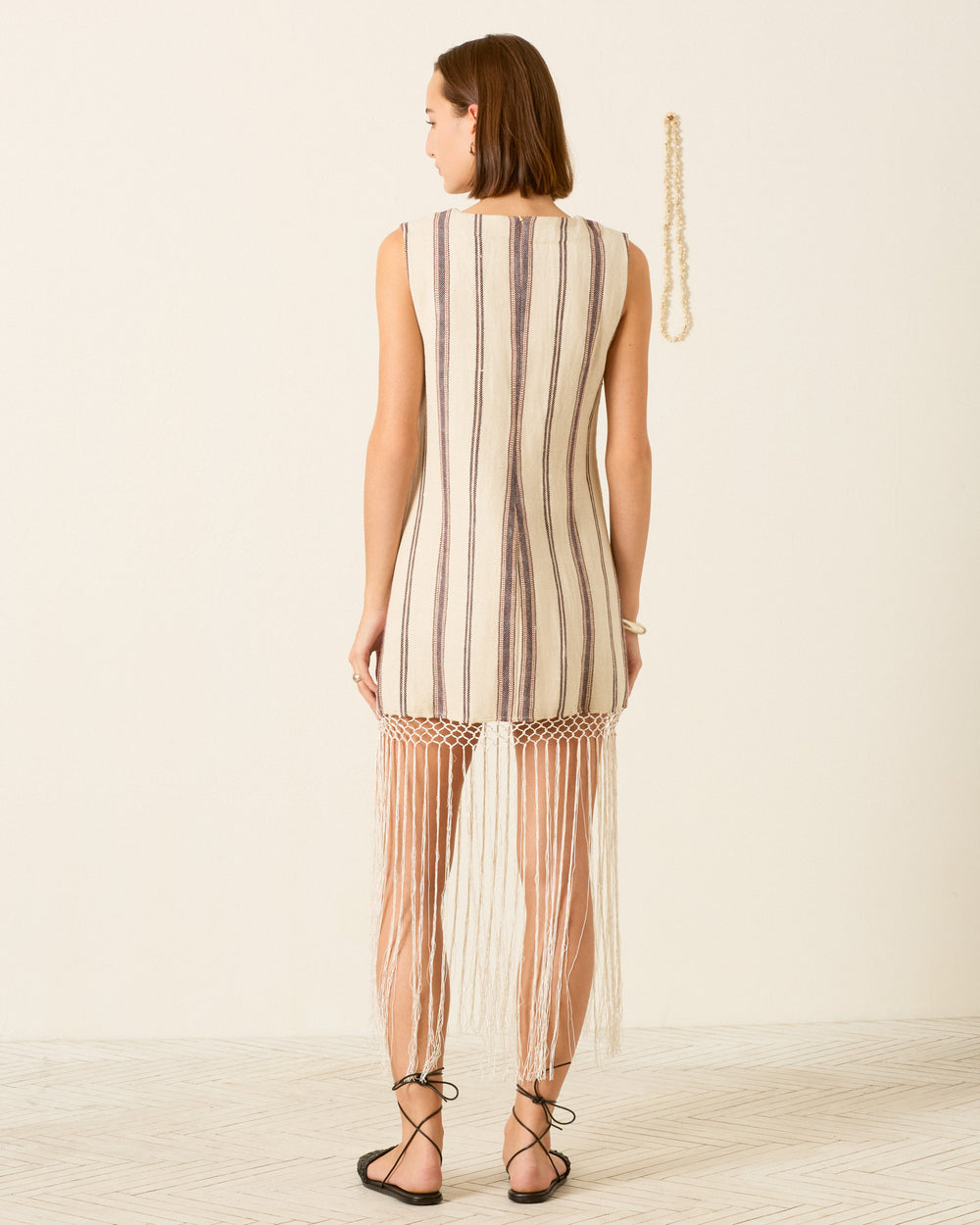 A woman faces away, wearing the Vita Bark Stripe Dress—a sleeveless shift in a linen-cotton blend with stripes and long fringe at the hem—paired with black sandals. A beaded necklace hangs on the wall behind her.
