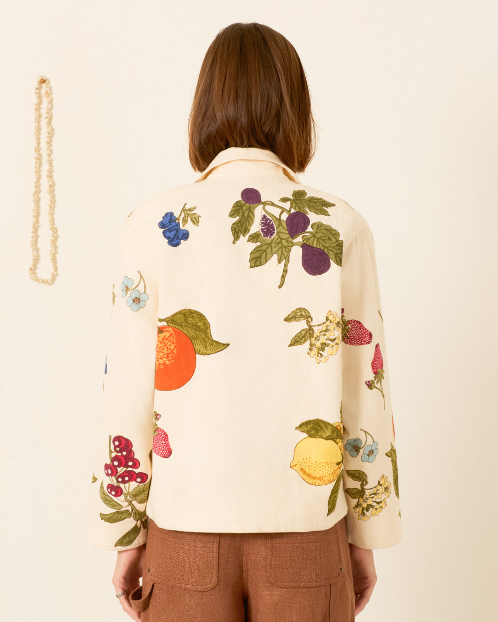 A person stands with their back to the camera, wearing the Western Nine Fruit Jacket—an oversized khadi cotton piece in cream, adorned with colorful hand-block-printed fruit motifs—paired with brown pants. A beaded necklace hangs on the wall to the left.