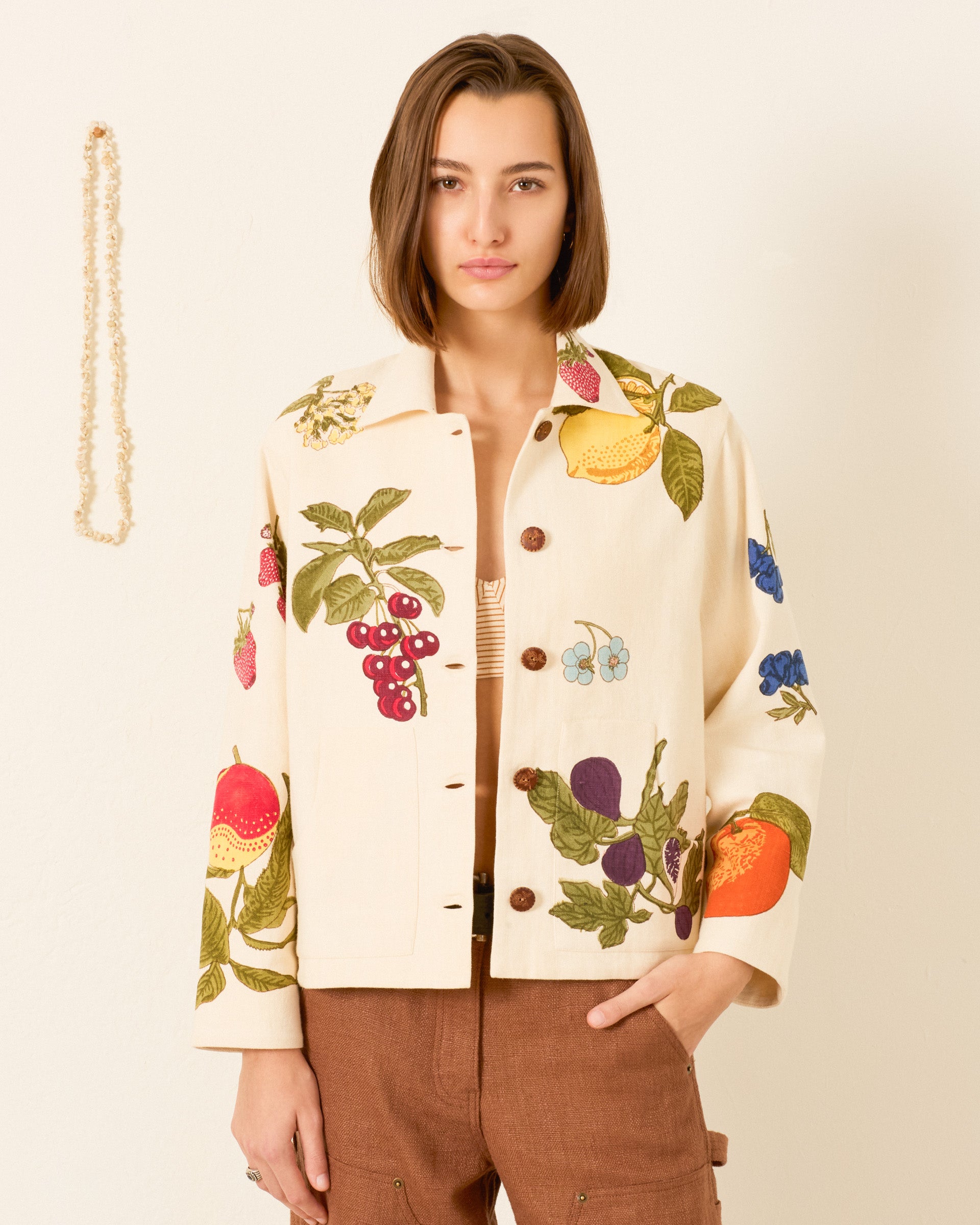A person wearing the oversized Western Nine Fruit Jacket, made from cream-colored Khadi cotton with colorful fruit prints, and brown pants stands against a plain wall.