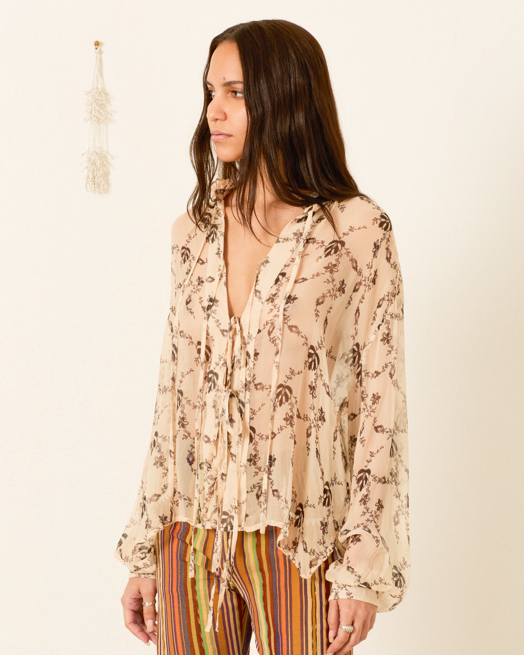 A woman models the Willa Shadow Lace Shirt, a sheer floral blouse with lace-inspired details and a tie-front, paired with multicolored vertical striped pants against a plain wall.