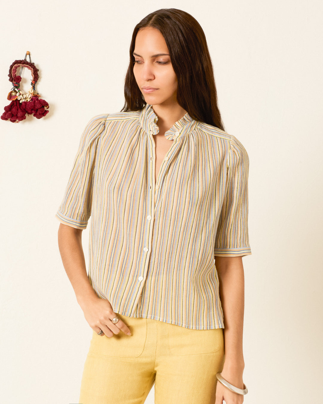 A woman wearing the Winn Sandlilt Stripe Shirt with puff shoulders and yellow pants stands against a plain wall with a decorative item hanging beside her.