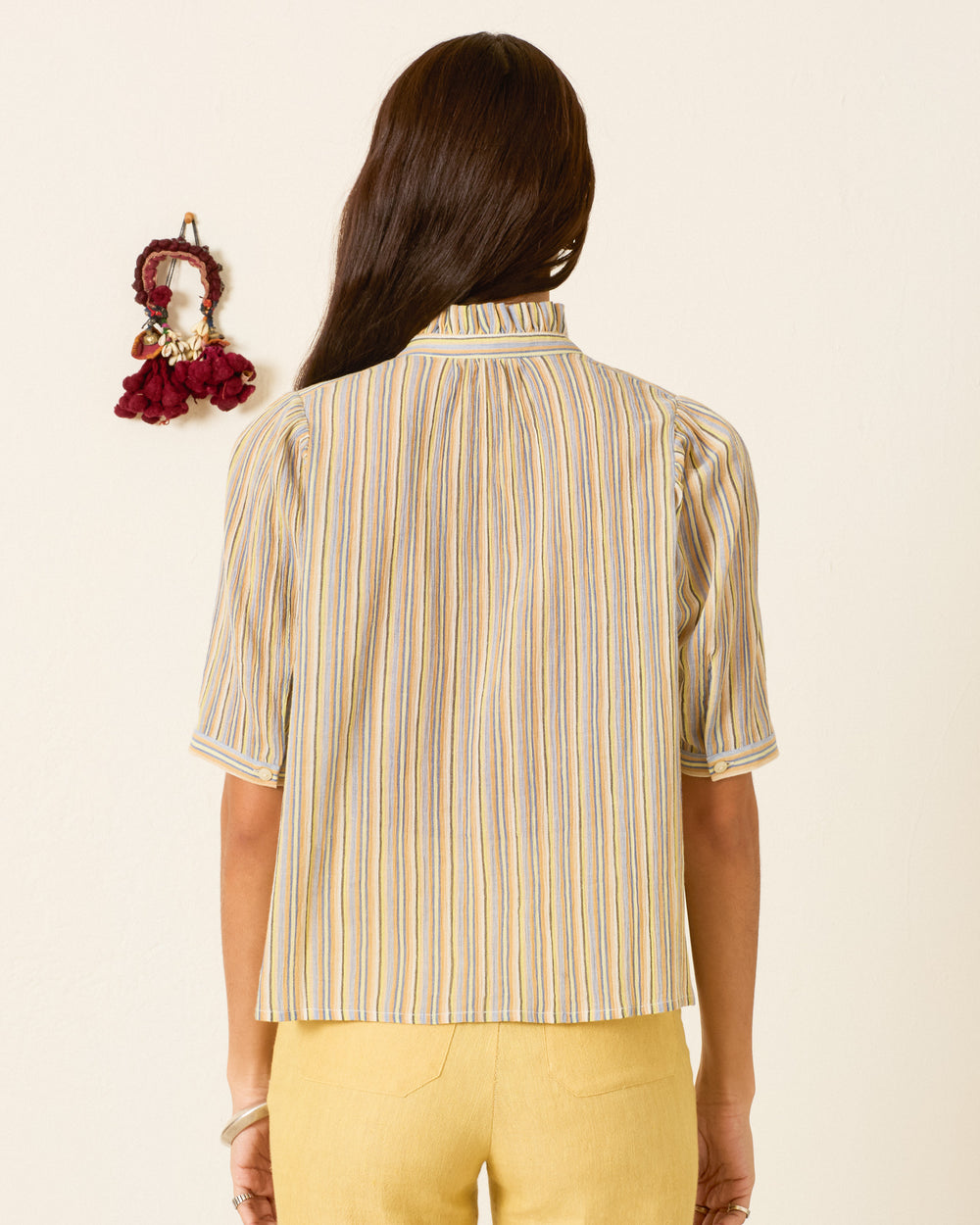 A woman stands facing away, wearing the Winn Sandlilt Stripe Shirt in yellow and matching pants, with a decorative wall hanging to the left.
