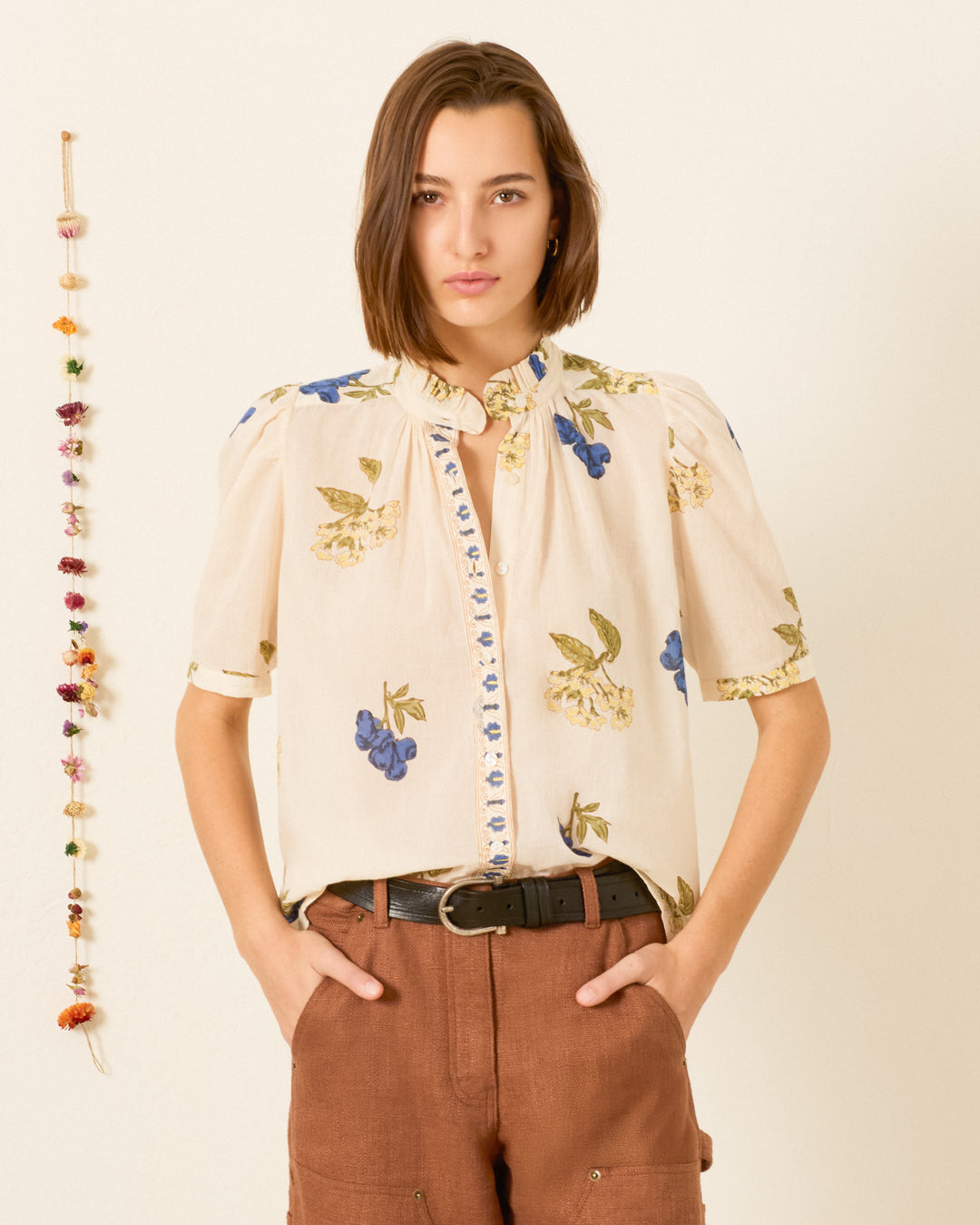 A woman stands with her hands in her pockets against a plain background, wearing the Winnie Blueberry Flora Shirt, a short-sleeved, hand-block-printed floral-print cotton voile top with a ruffled collar, paired with brown pants. A string of small decorations hangs on the wall behind her.