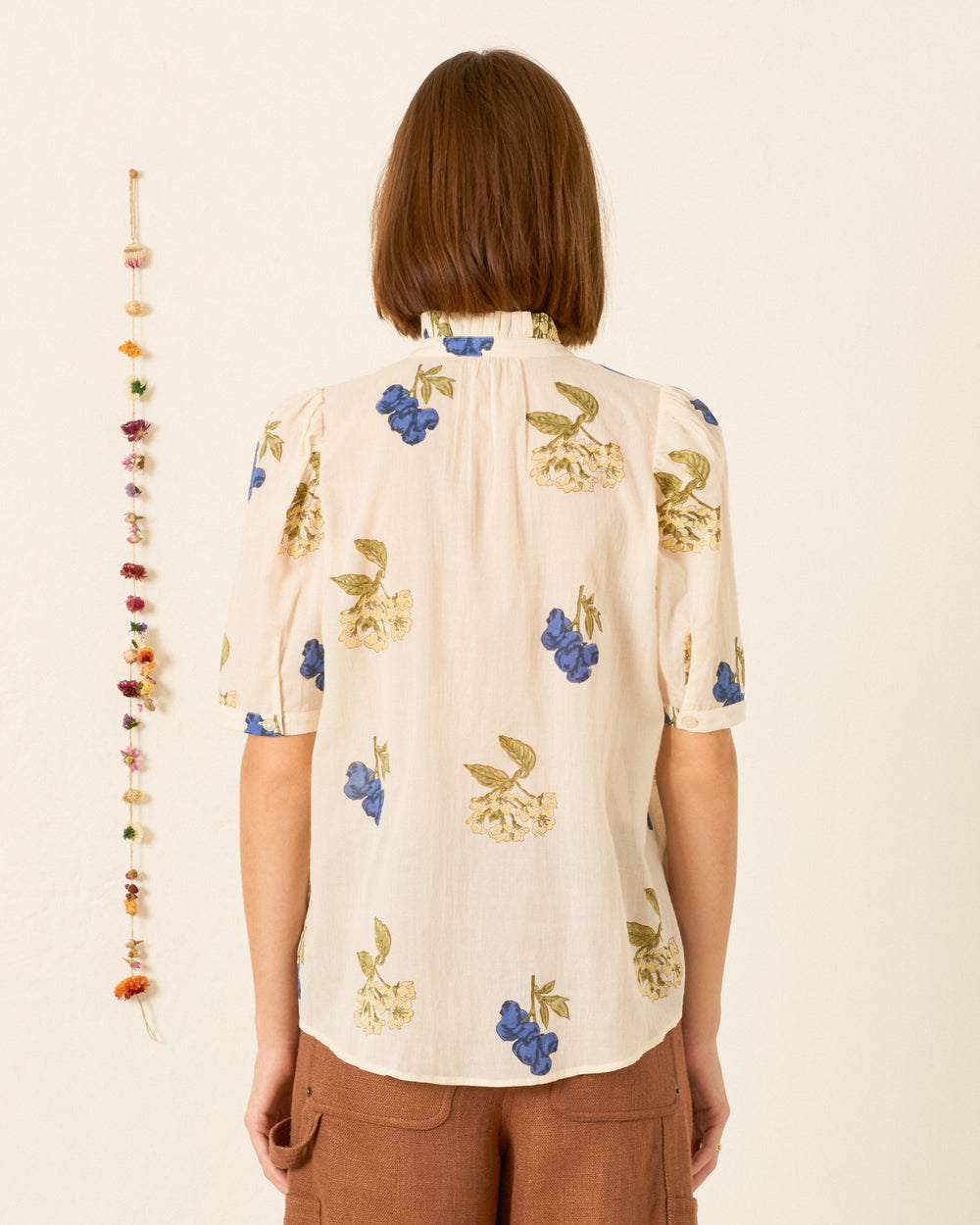 A person with chin-length brown hair faces away, wearing the short-sleeved Winnie Blueberry Flora Shirt, which features hand-block-printed floral designs and a ruffled collar, paired with brown shorts. Colorful beaded wall decor hangs to the left against a plain cream-colored background.
