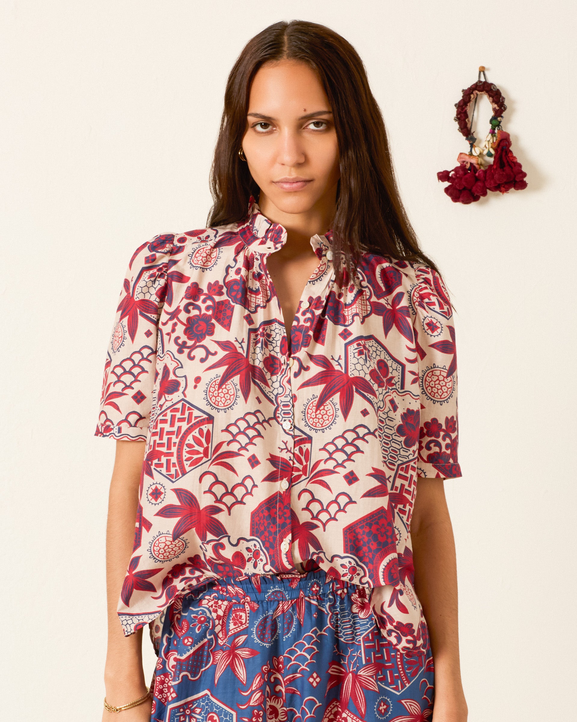 A woman stands indoors wearing the Winnie Crimson Hive Shirt, a patterned short-sleeve blouse with red and blue designs and subtle puff shoulders. A maroon accessory hangs on the wall behind her, and she pairs it with a blue skirt.