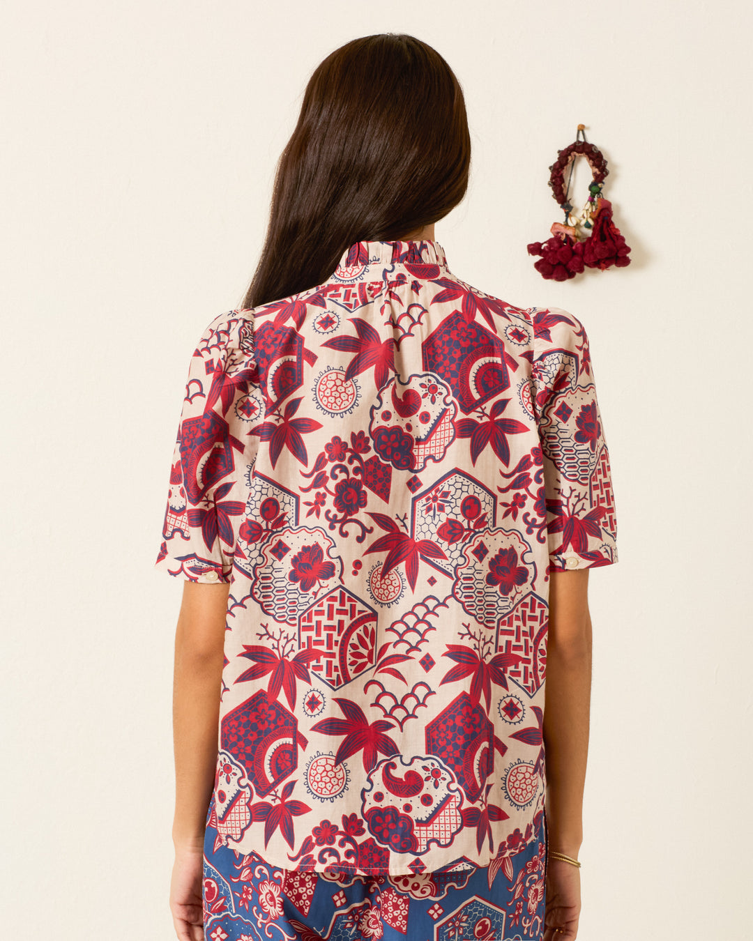 A woman with long brown hair faces away, wearing the Winnie Crimson Hive Shirt—a short-sleeve top featuring a red and pink geometric and floral pattern with subtle puff shoulders. A wall decor item hangs on the beige wall beside her.