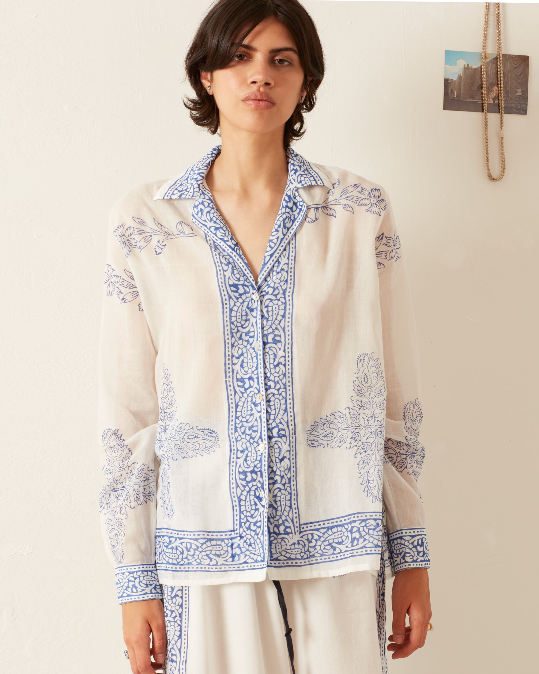 A person is dressed in a relaxed fit Patti Criss Cross Shirt, featuring intricate blue floral patterns against a white backdrop, standing against a light-colored wall. In the background, a small photograph and a chain hang on the wall, enhancing the serene ambiance.