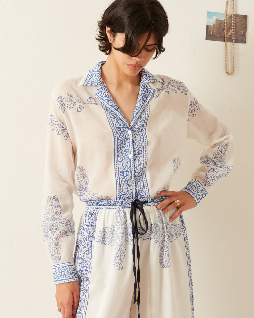 Dressed in the relaxed-fit Patti Criss Cross Shirt, a person wears a long-sleeved white outfit adorned with blue floral patterns. The V-neck shirt is paired with matching pants as they stand looking down against a plain white background, where a small photograph and a necklace hang on the wall behind them.