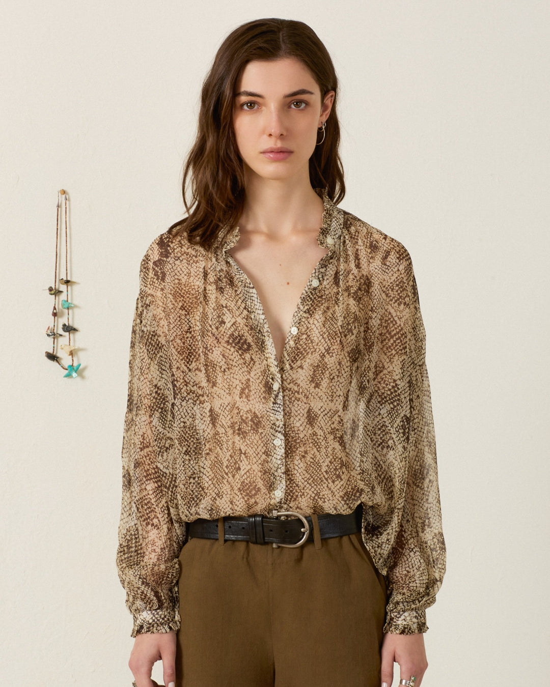 A woman stands indoors wearing the Poet Lemon Snakeskin Blouse, a vintage-inspired brown shirt with a ruffled collar, paired with brown pants; a necklace hangs on the wall beside her.