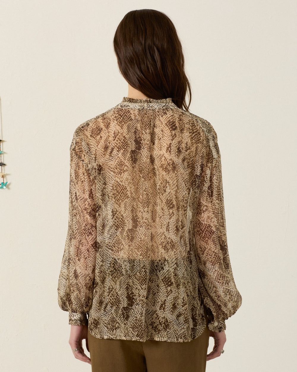 A person with long brown hair is standing facing away, wearing the Poet Lemon Snakeskin Blouse—a vintage-inspired, sheer snakeskin-print shirt with long sleeves—paired with brown pants against a plain background.
