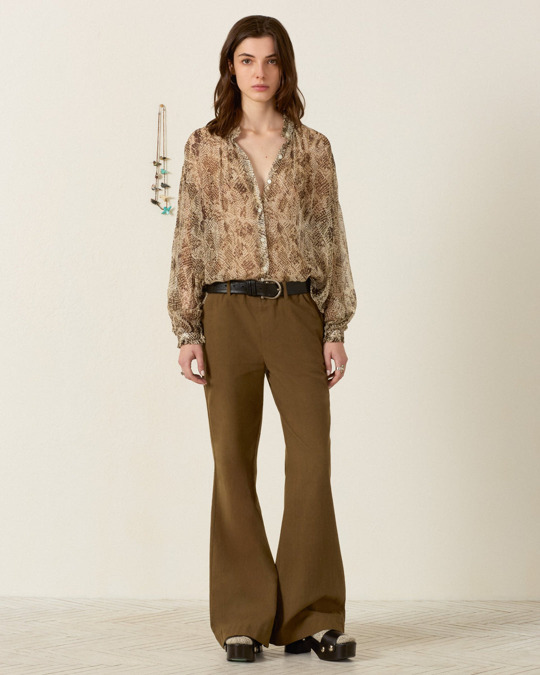 A woman stands against a plain wall, wearing the Poet Lemon Snakeskin Blouse—a vintage-inspired brown patterned blouse with a ruffled collar—paired with wide-leg brown pants and black platform shoes. A hanging necklace is visible on the wall behind her.