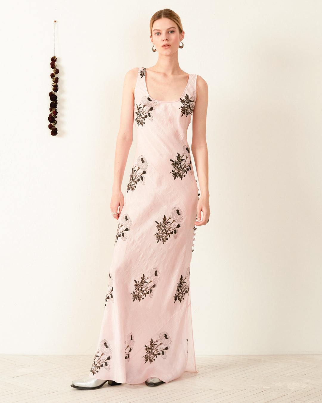 A person is elegantly dressed in the Adele Anemone Silk Dress, a bias-cut slip featuring black and gray floral designs. The long, sleeveless pink dress beautifully enhances the room's simple decor, adding a touch of grace to the plain background with its intricate patterns.