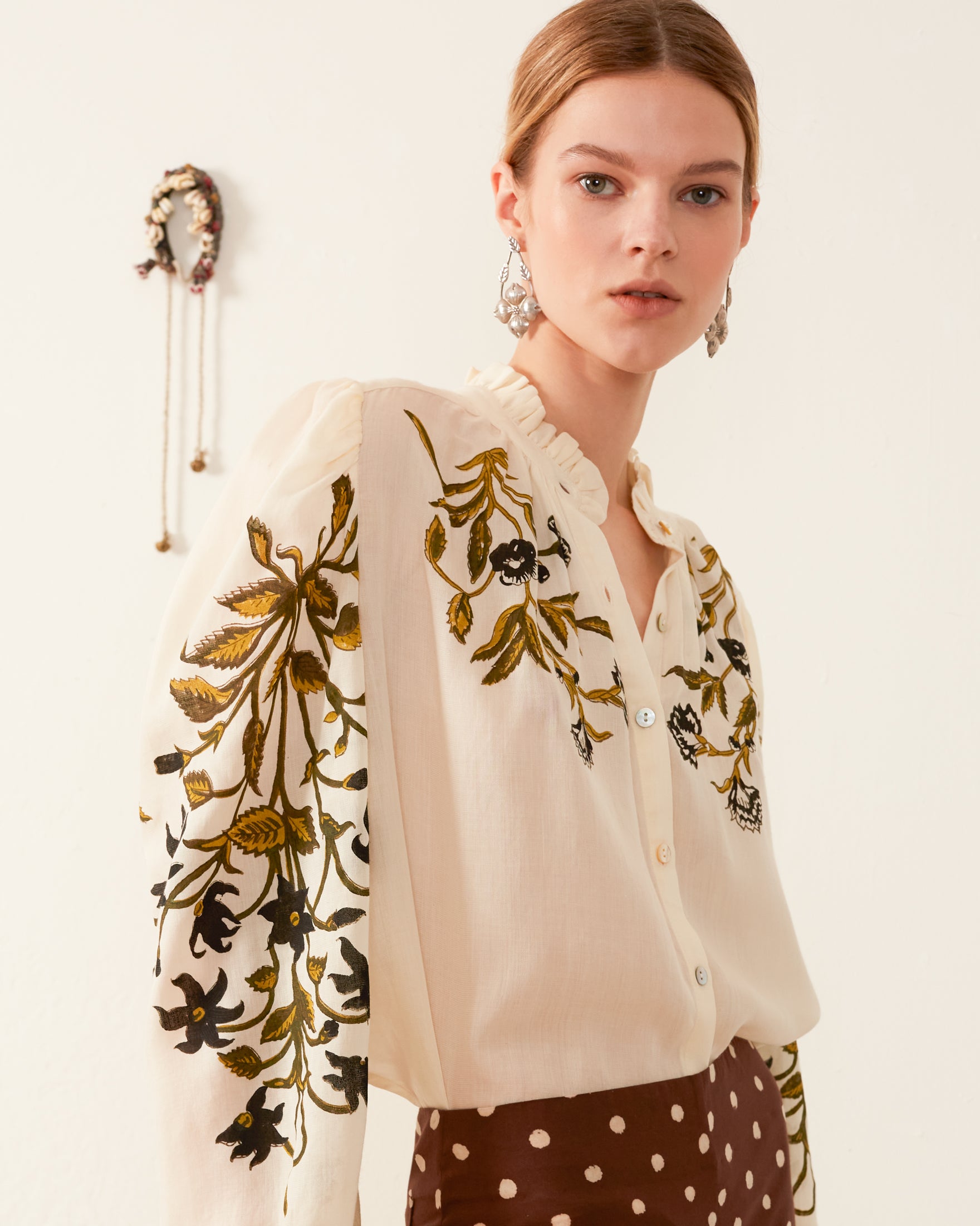 A woman stands against a light background wearing the Annabel Olive Lily Valley Shirt, which showcases a cream color with floral motifs and a ruffled collar for an elegant touch. She pairs it with a brown polka dot skirt and accessorizes with dangling earrings. A decorative hanging item can be seen on the wall.