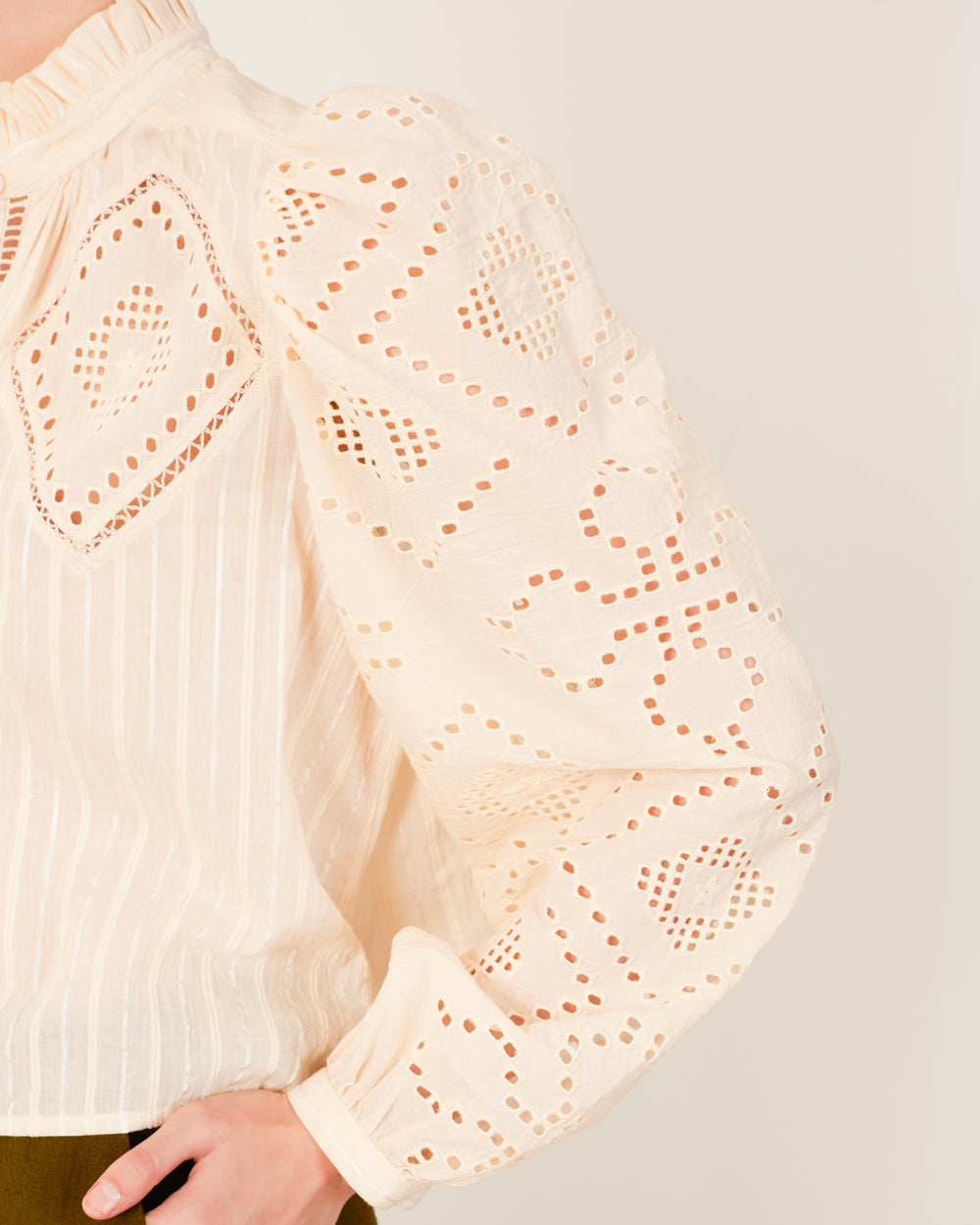 A close-up showcases someone wearing the Annabel Pearl Eyelet Shirt, a cream-colored blouse featuring puffed sleeves and a ruffled collar. The fabric is adorned with hand embroidery and intricate cut-out patterns, providing a texture similar to that of an eyelet design. The person is posed against a plain background.