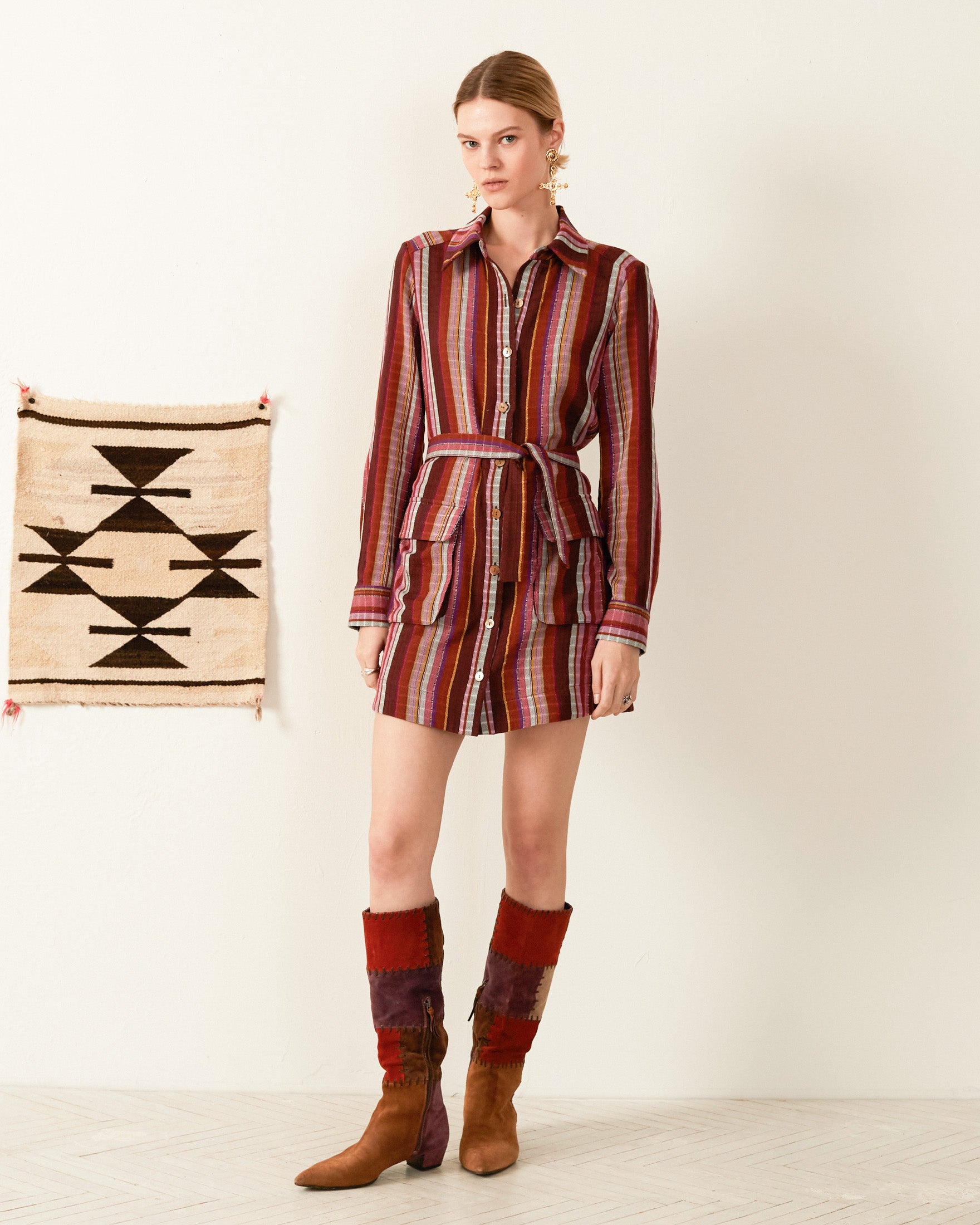 A person stands against a light wall, adorned in the Arlo Truffle Berry Dress paired with knee-high brown boots. Beside them, a hand-loomed Guatemalan ikat tapestry featuring geometric patterns hangs gracefully. The floor is light wood.