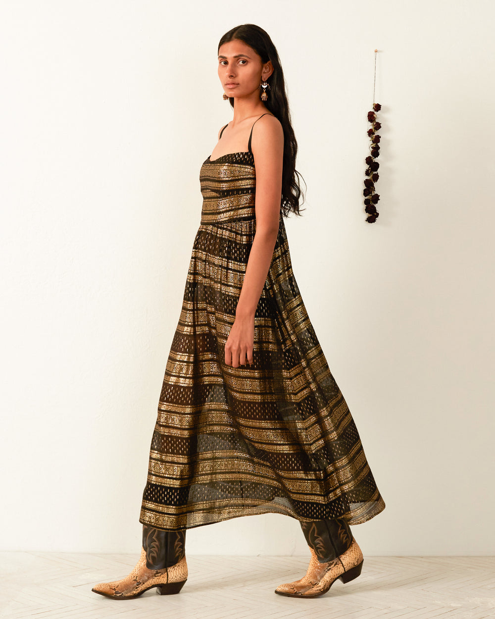 A person with long dark hair stands against a plain white wall, showcasing the Autumn Gilded Lily Dress and brown boots. They are facing to the left with earrings visible, and a string of decorative elements hangs on the wall nearby.