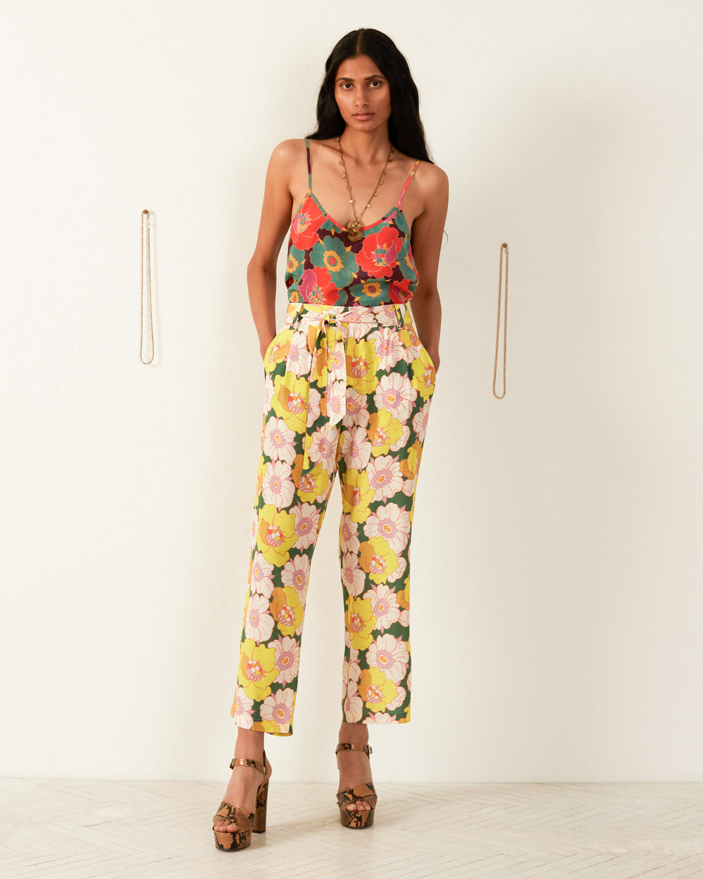 A woman with long dark hair stands against a plain background, wearing a floral Liberty-print camisole and the Colette Citrus Blossom Silk Pant featuring red, green, and yellow blooms. She completes her look with platform heels and casually places her hands in her pockets.