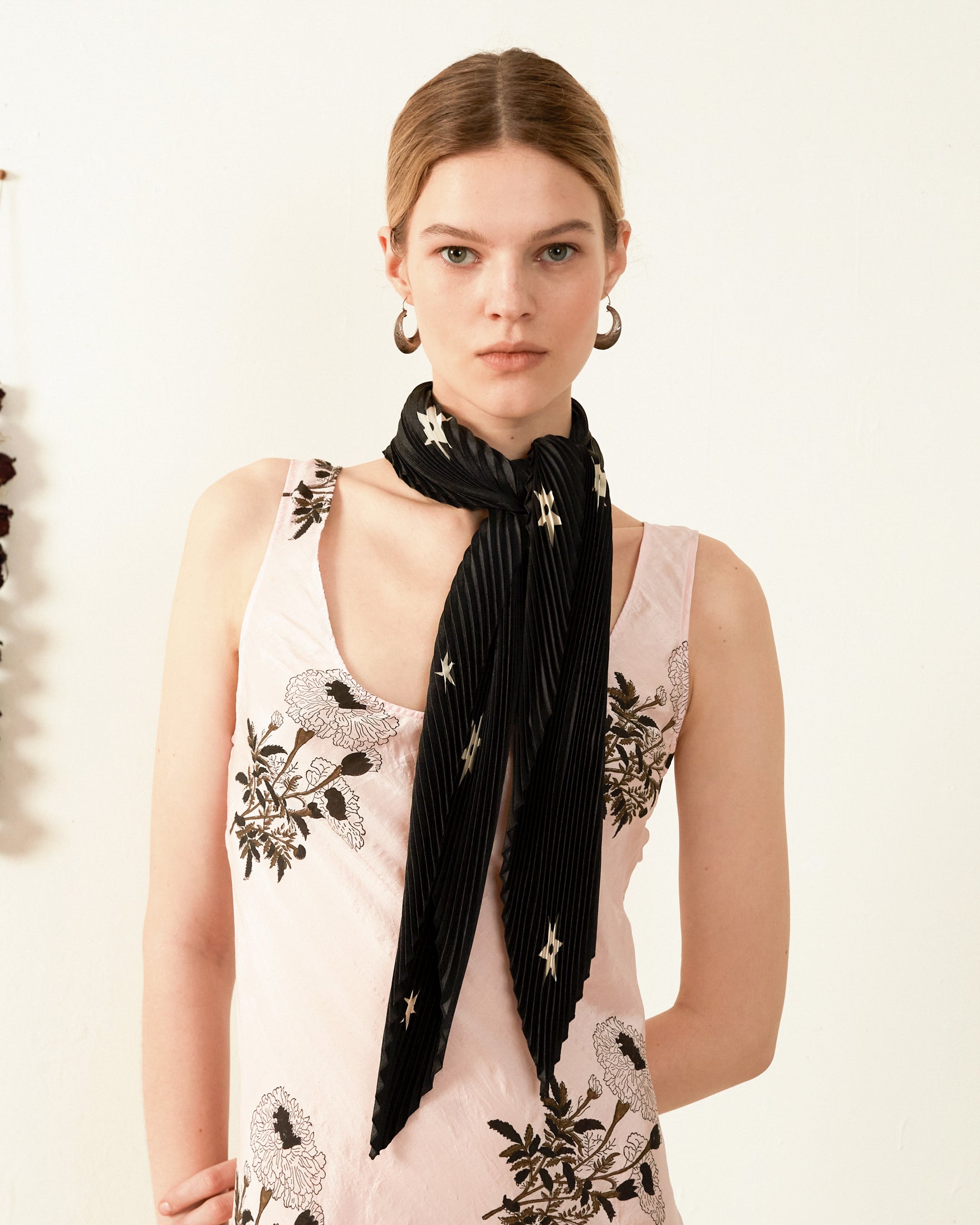 A person with light brown hair is wearing a sleeveless, light-colored dress adorned with black floral embroidery. An Estella Noir Plisse Scarf, featuring star patterns, gracefully adorns their neck as they stand against a plain, off-white background.