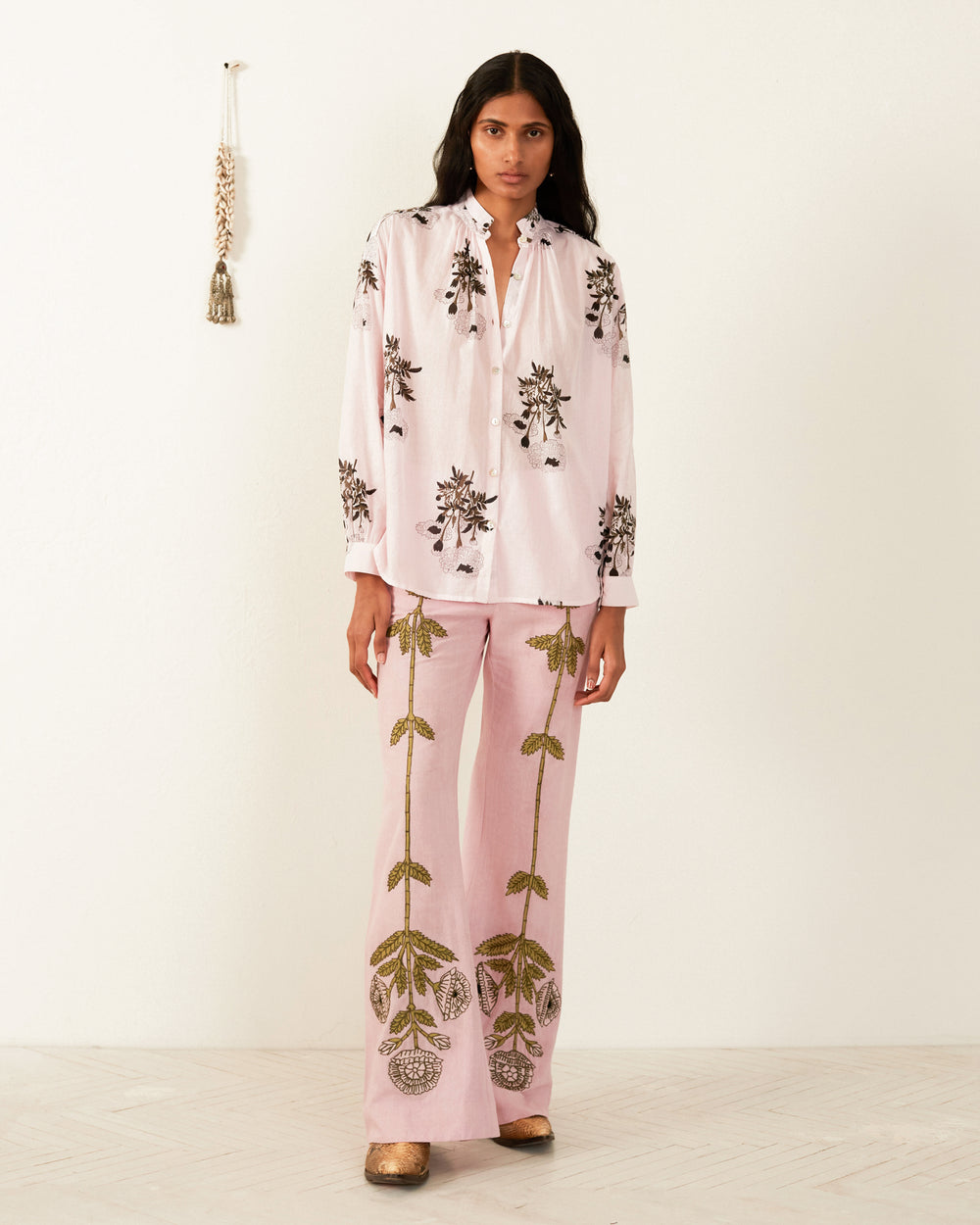 A person stands against an off-white wall, wearing the Kiki Anemone Bloom Shirt, featuring light pink floral prints with intricate black designs. The gold floral patterns adorn the wide-leg pants made from hand-woven textile. They are also wearing gold shoes, complemented by a tassel decoration hanging on the wall.