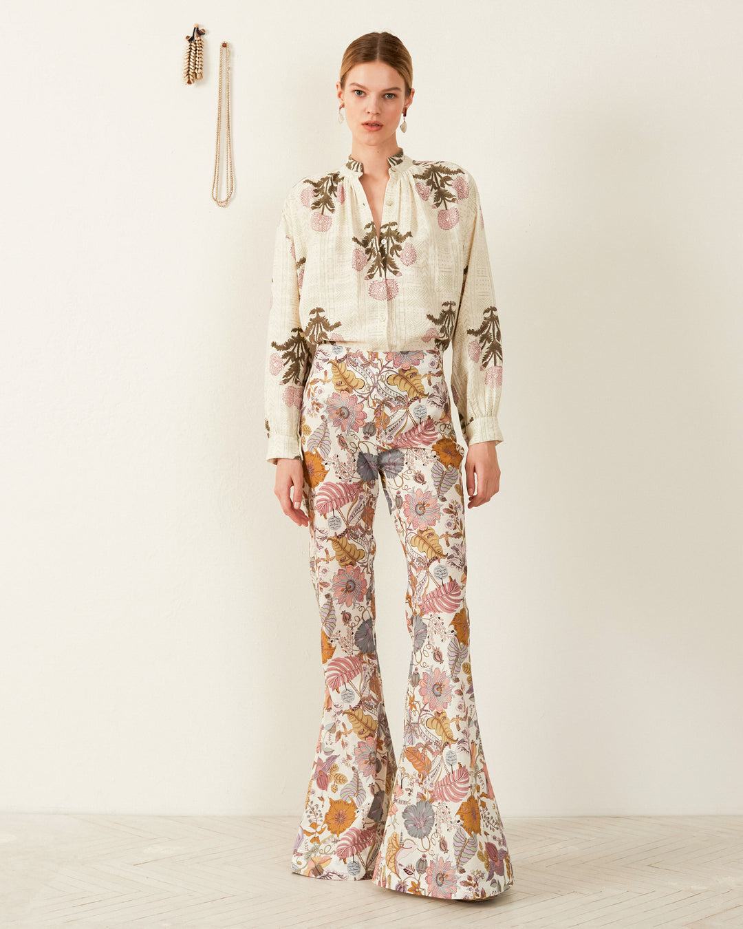 A person wearing a Kiki Blush Triple Bloom Shirt and matching wide-leg pants, both hand-loomed in earthy tones with intricate floral patterns, stands against a white wall. A necklace and a bracelet are hung on the wall to the left.