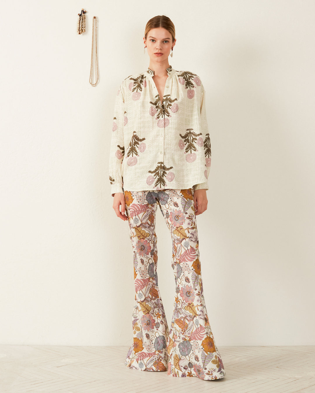 A person stands against a light background, wearing the Kiki Blush Triple Bloom Shirt—a white blouse adorned with floral patterns—and multicolored bell-bottom pants. This hand-loomed cotton shirt adds a unique touch, complemented by a necklace and bracelet hanging on the wall, which enhance the vintage and eclectic style.