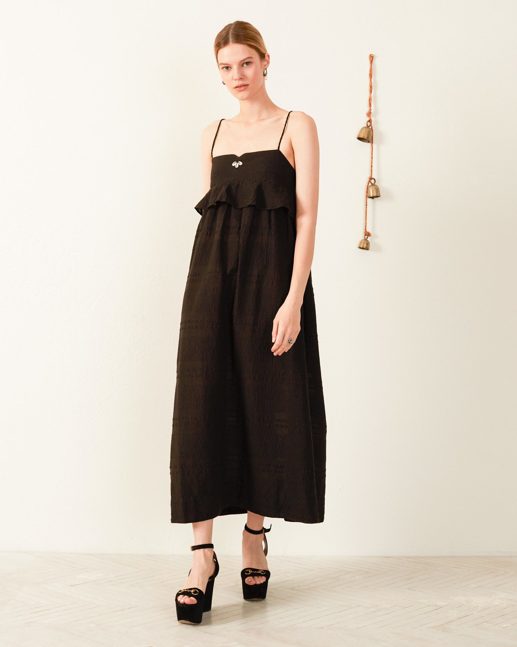 A person wearing the Martina Noir Seersucker Dress, a black sleeveless piece with ruffled detailing and an empire waist, along with black platform heels, stands in a minimalist room. A decorative hanging embellished with cowrie shells and bells adorns the wall behind them.