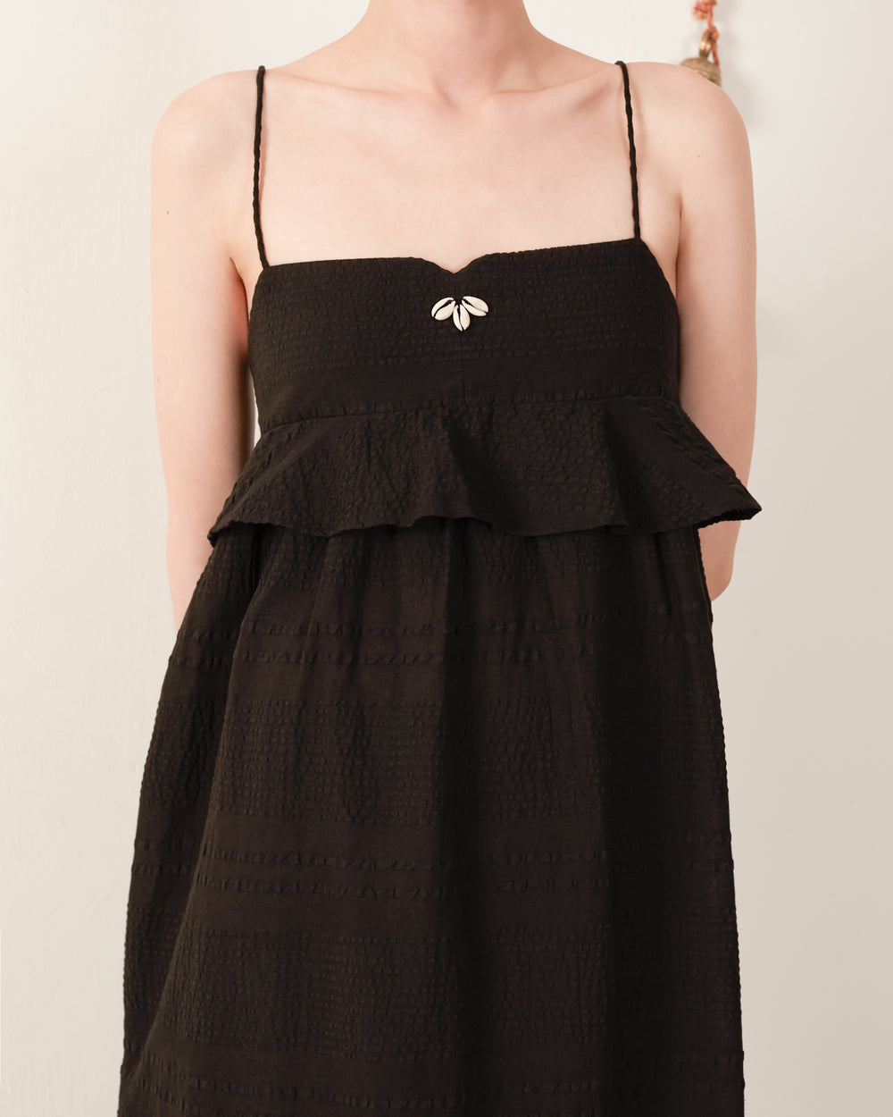 The scene is graced by a figure wearing the Martina Noir Seersucker Dress, a sleeveless black piece with thin straps and an empire waist. The ruffled top section showcases a small flower ornament at the center of the neckline, all set against a plain, light-colored background.