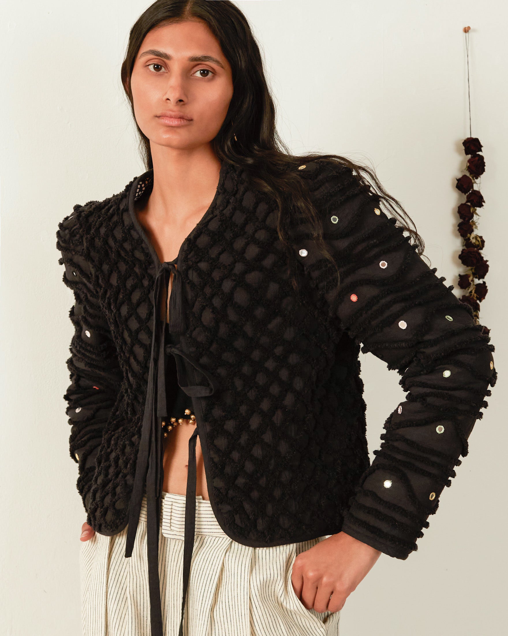 A person with long dark hair wears the Pasha Onyx Shag Jacket, a hand-embroidered, cropped black jacket adorned with sequins, over a black top featuring a tie-front design. They pair it with light-colored striped pants. The backdrop is simple, showcasing a Moroccan Handira blanket hanging to the side.