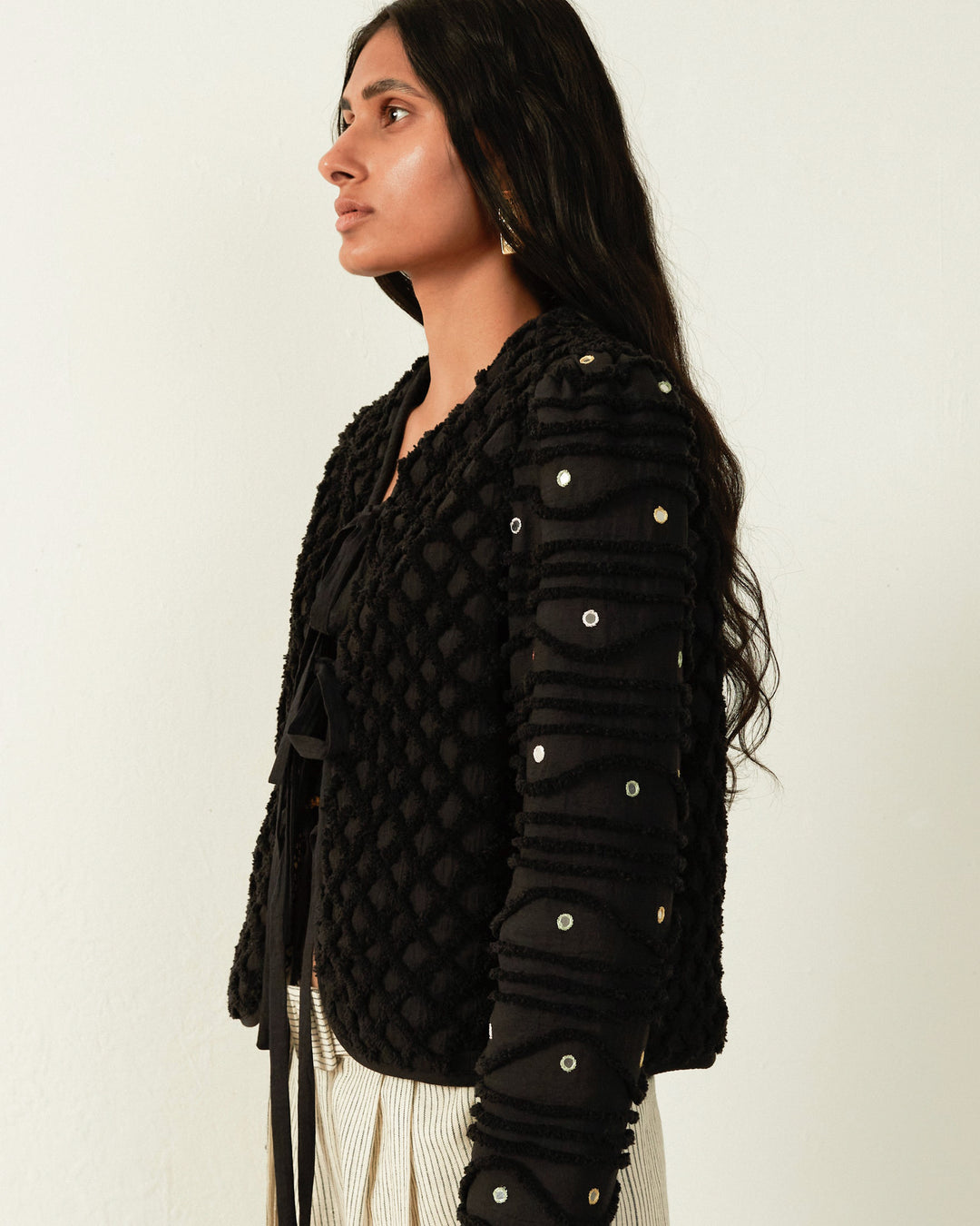 A person with long dark hair wearing the Pasha Onyx Shag Jacket, adorned with circular embellishments, stands in profile against a light background, reminiscent of the intricate artistry seen in Moroccan Handira blankets.