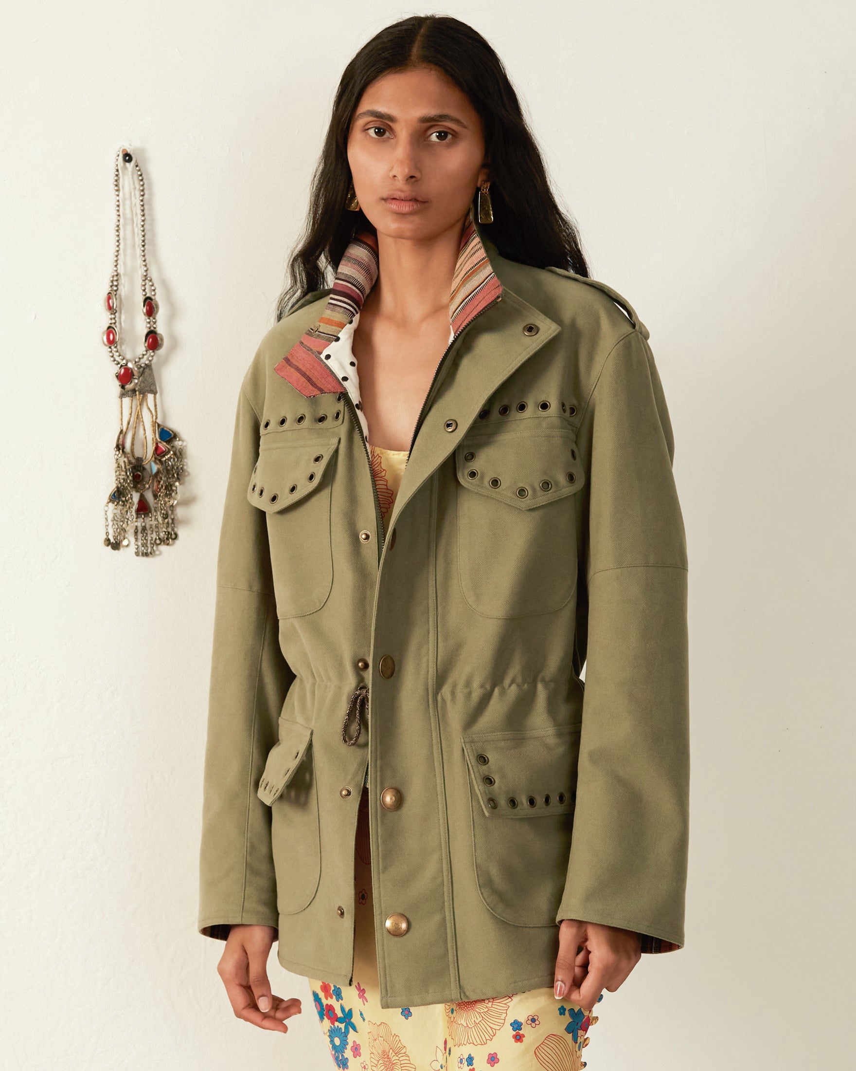 A person with long dark hair wears the Porter Eucalyptus Moleskin Jacket, an army green utility piece made from Japanese brushed cotton, featuring decorative studs and multiple pockets. They stand against a plain wall, with a beaded necklace hanging in the background.