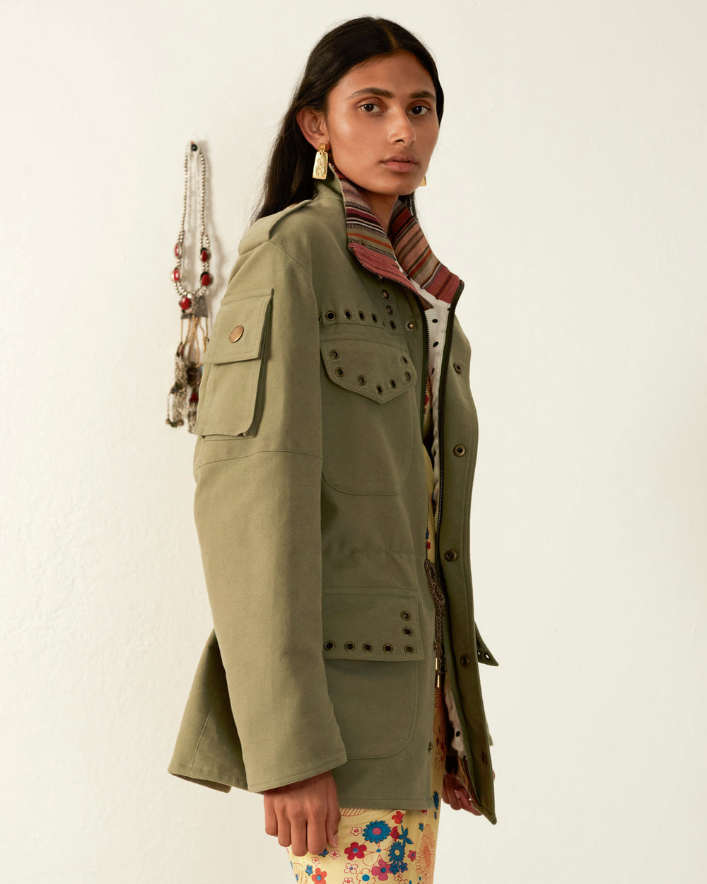 A person wearing a Porter Eucalyptus Moleskin Jacket with brass studs over a colorful, patterned dress stands against a white wall. A beaded necklace hangs on the wall in the background. They have long, dark hair and are looking at the camera.