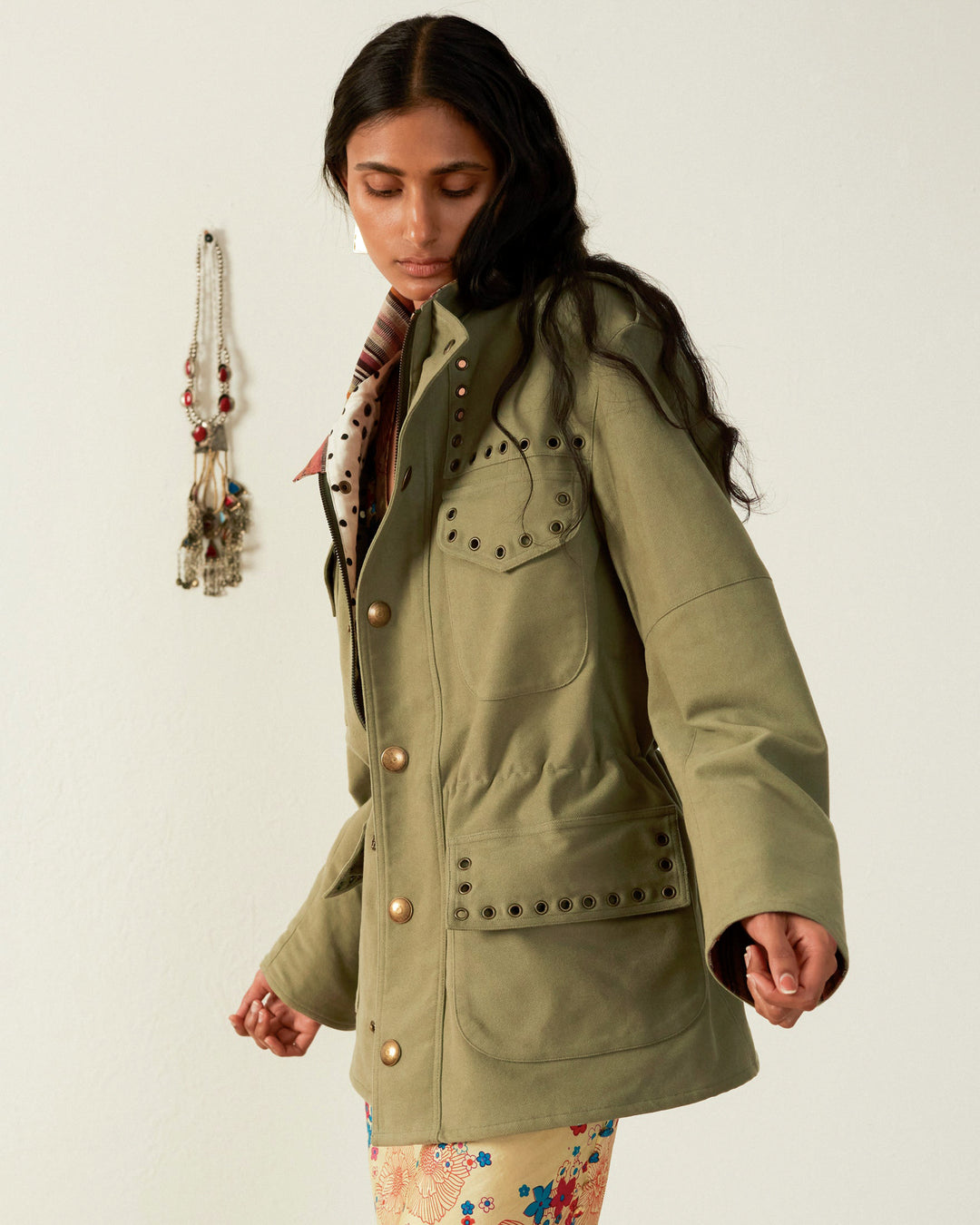 A person with long dark hair wears the Porter Eucalyptus Moleskin Jacket, crafted from olive green Japanese brushed cotton and accented with decorative buttons and eyelets. They gaze downwards in a light-colored room that features a small wall ornament in the background.