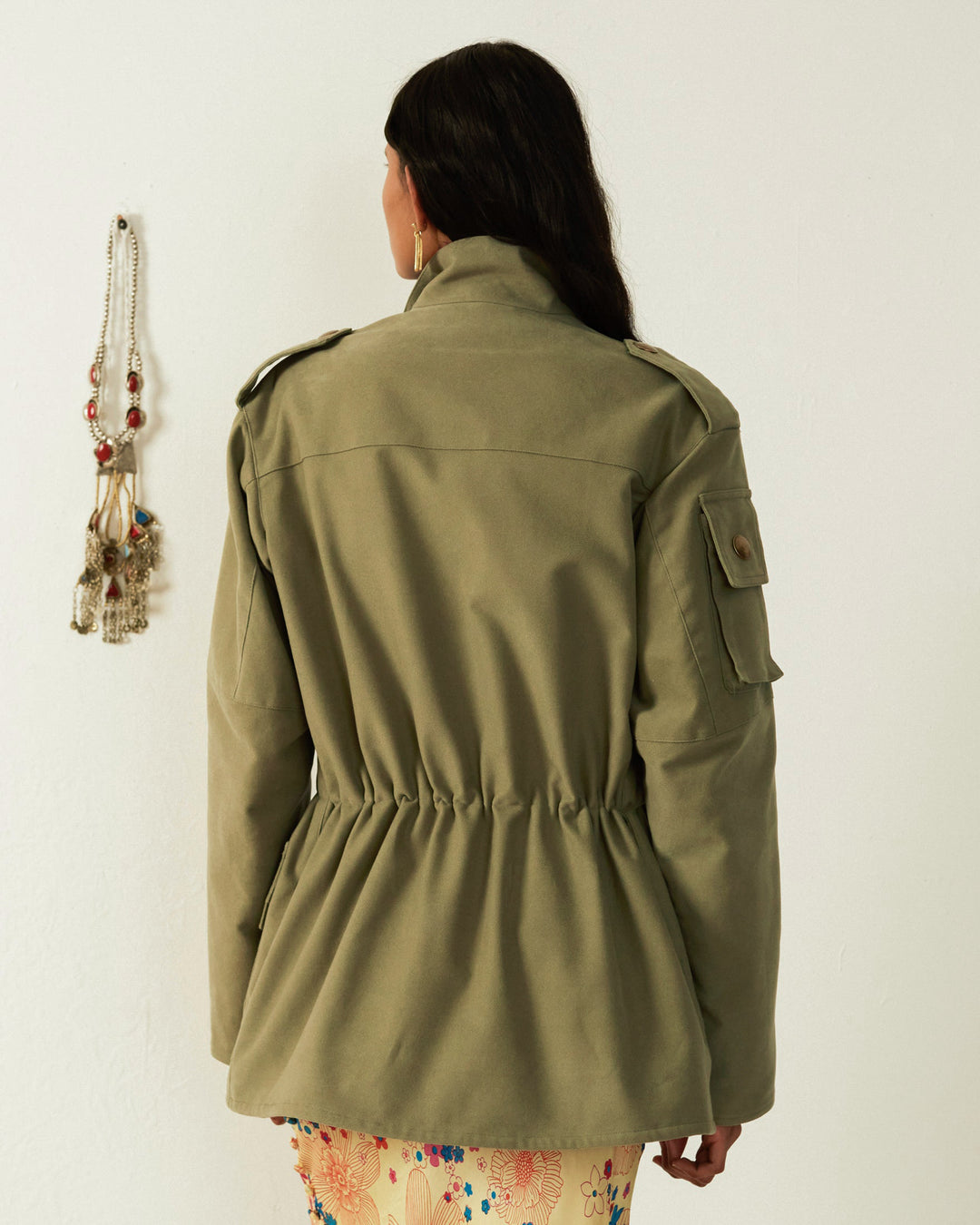 Rear view of a person with long dark hair wearing the Porter Eucalyptus Moleskin Jacket in army green, featuring a cinched waist, alongside patterned pants. A beaded accessory is hanging on the wall beside them.