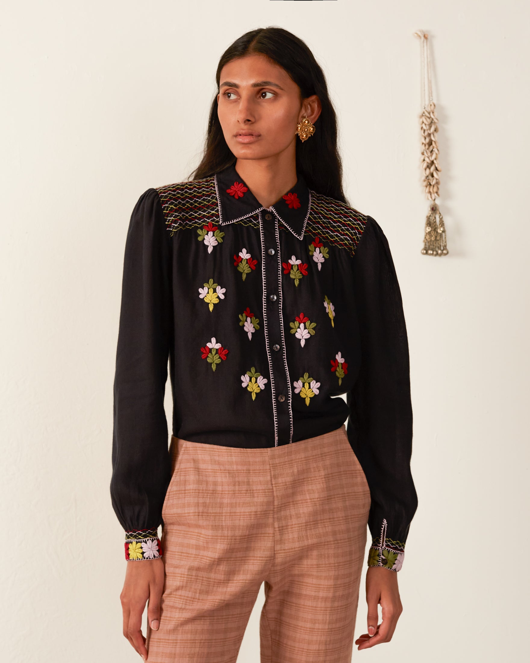 A person with long dark hair wearing the Priscilla Smoke Smocked Blouse featuring colorful floral embroidery and light brown plaid pants stands against a plain white wall. A small decorative item hangs on the wall.