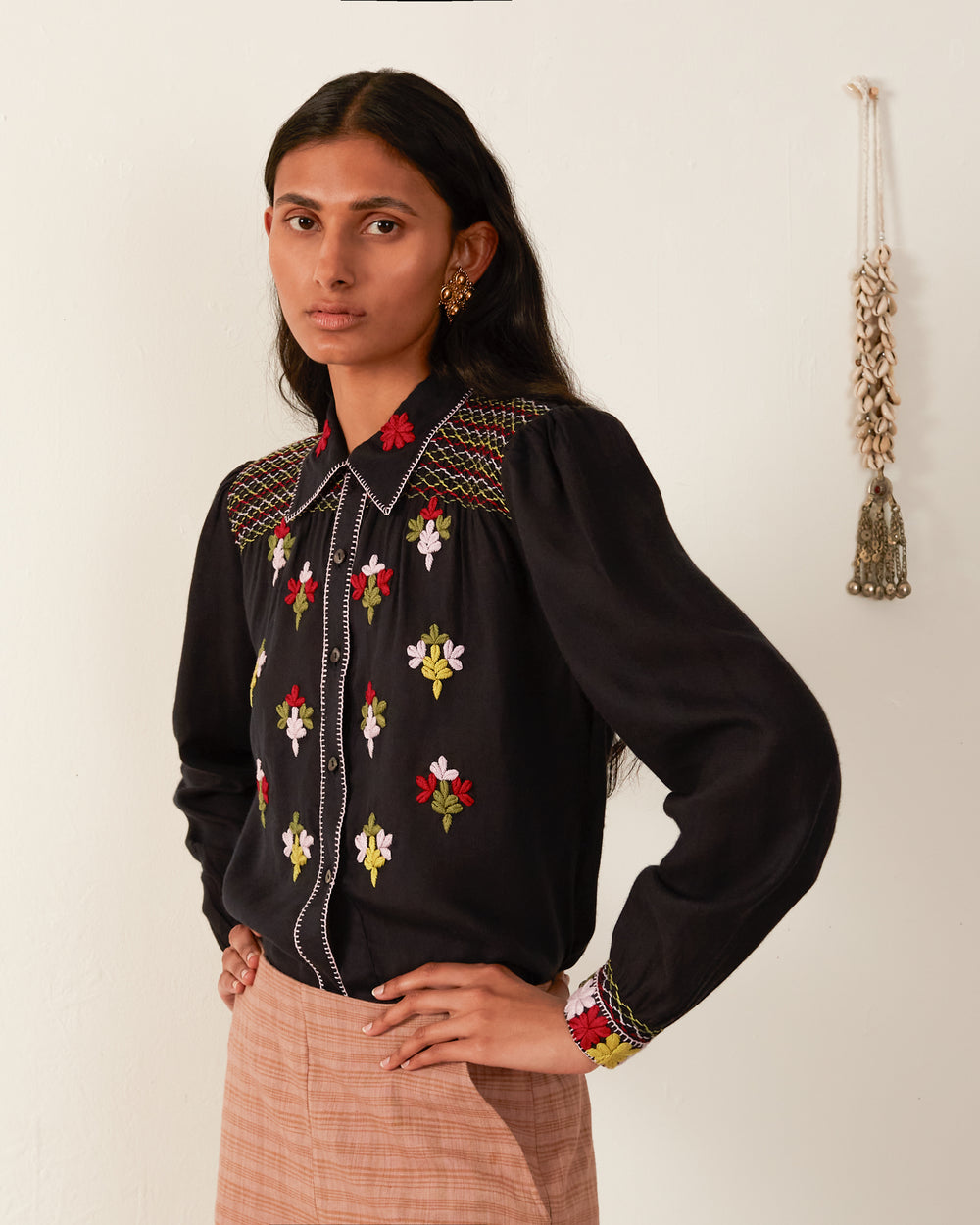 A person with long dark hair stands confidently, wearing the Priscilla Smoke Smocked Blouse, featuring colorful floral embroidery and a hand-smocked placket. They pair it with a light brown skirt. A decorative tassel hangs on the wall in the background.