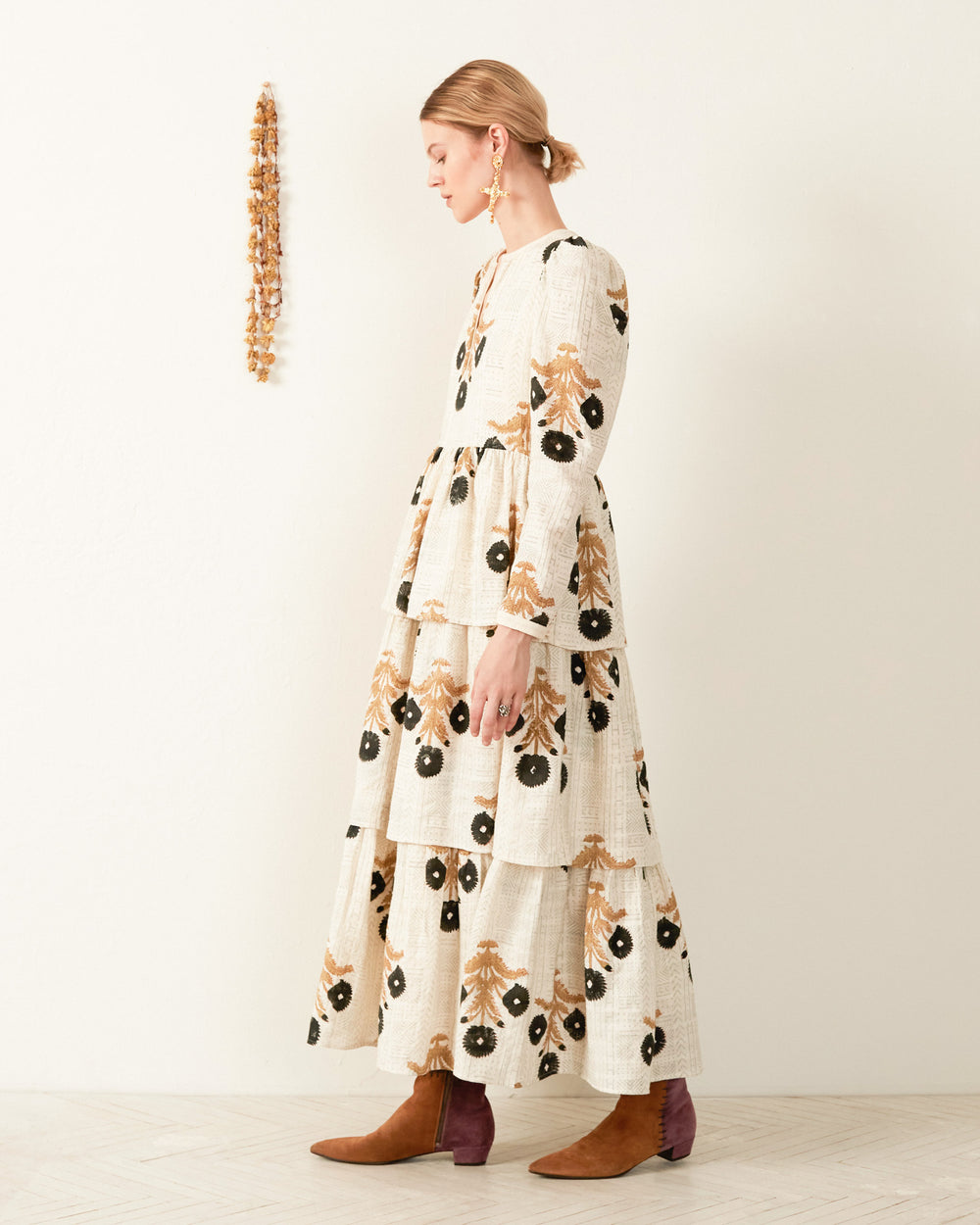 A person stands in profile, showcasing the Tallulah Noir Triple Bloom Dress, a long, tiered outfit with a striking block-printed floral design in beige, brown, and black. They have short hair and are paired with brown ankle boots. Nearby hangs a dried floral arrangement on the light-colored wall.