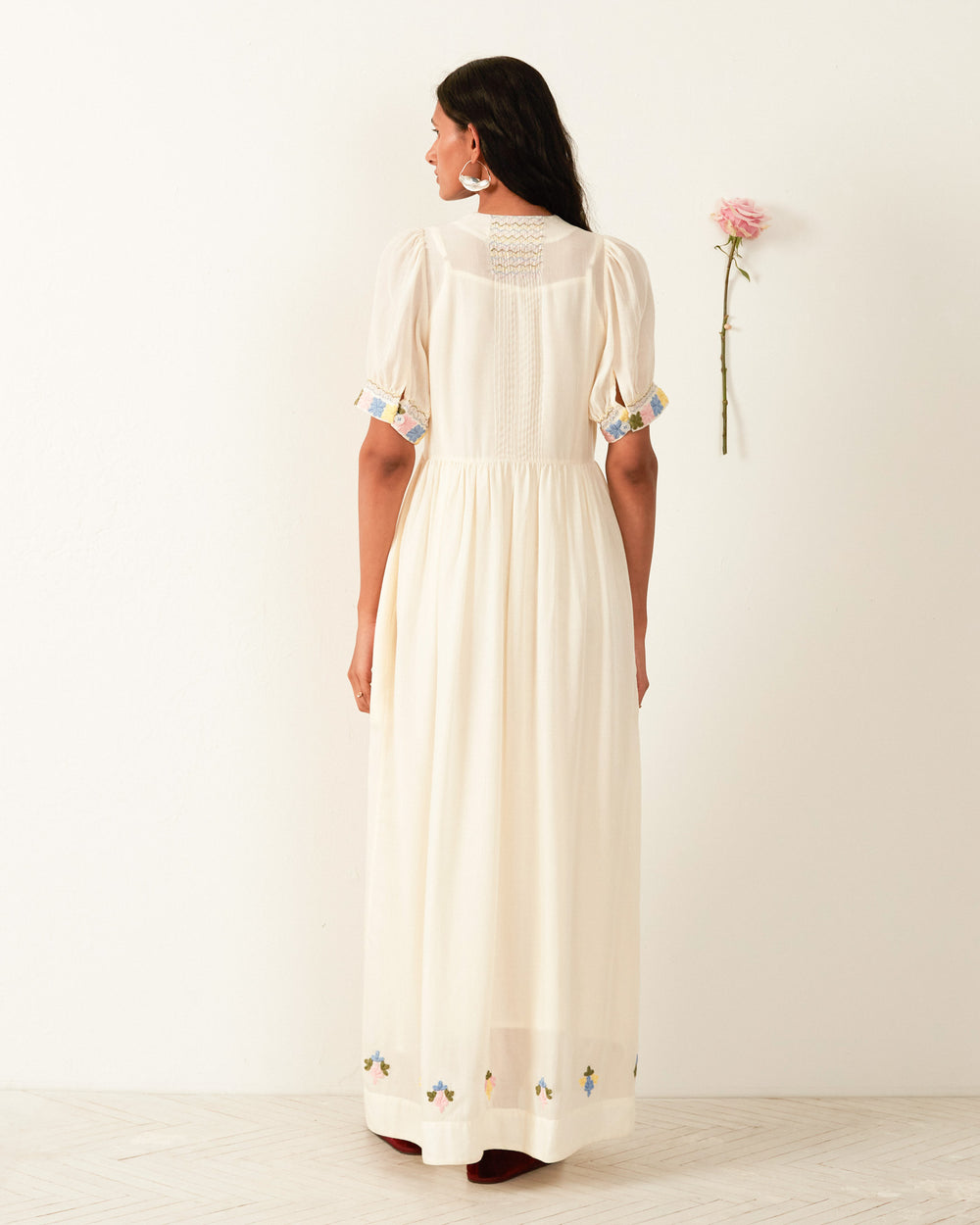 A woman is standing with her back to the camera, dressed in the Waverly Oyster Embroidered Dress, which features a long white design with vibrant floral embroidery on the sleeves and hem. Her long, dark hair cascades down as she faces a plain white wall adorned with a single pink flower.