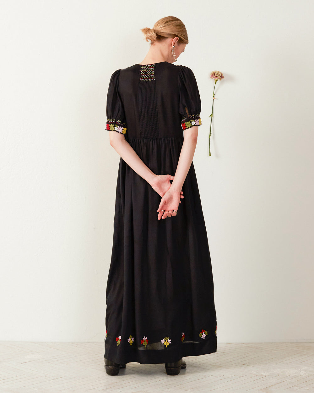 The individual stands with their back to the camera, donning the Waverly Smoke Embroidered Dress—a long black garment embellished with intricate floral embroidery on the sleeves and hem. They clasp their hands behind their back, and a single flower is pinned to the wall beside them.