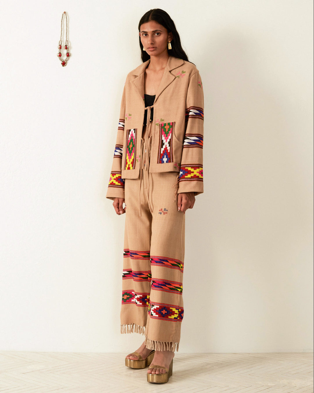 Against a white wall, a person wears a beige Western Dune Blanket Jacket featuring colorful geometric patterns on the sleeves, pockets, and pants. The oversized jacket is adorned with fringe details that highlight the hand-woven textiles. They pair it with platform sandals, while long beaded earrings hang nearby.