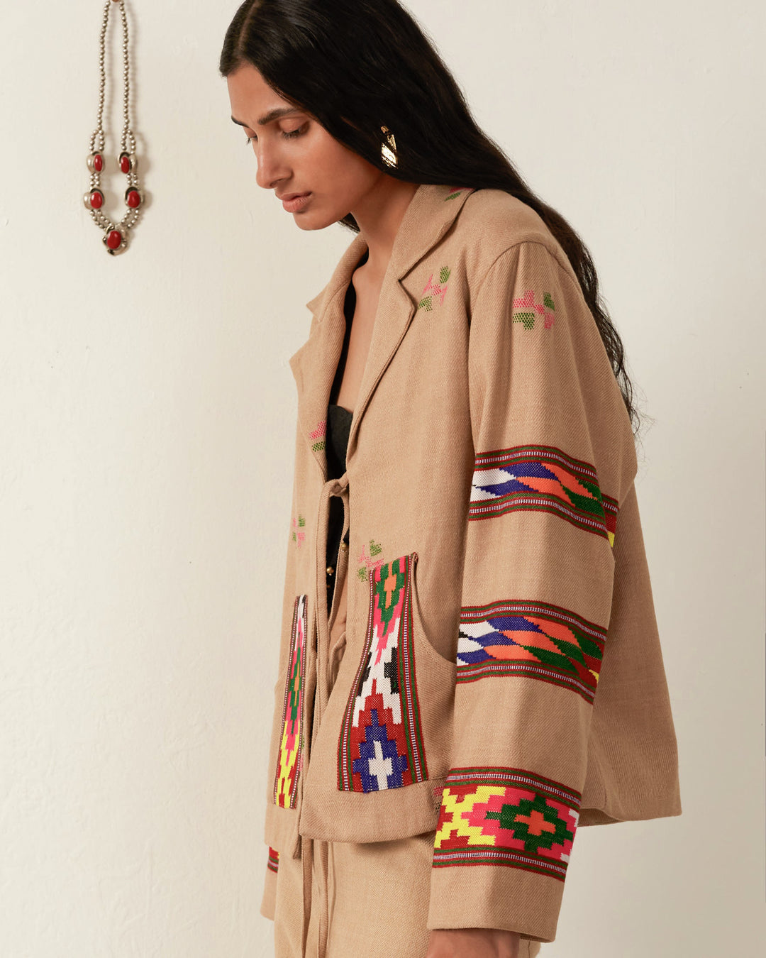 A woman with long dark hair is wearing a Western Dune Blanket Jacket, an oversized beige piece adorned with colorful geometric patterns on its sleeves and front. She is gazing downwards, while in the background, a decorative beaded necklace is displayed on the wall.