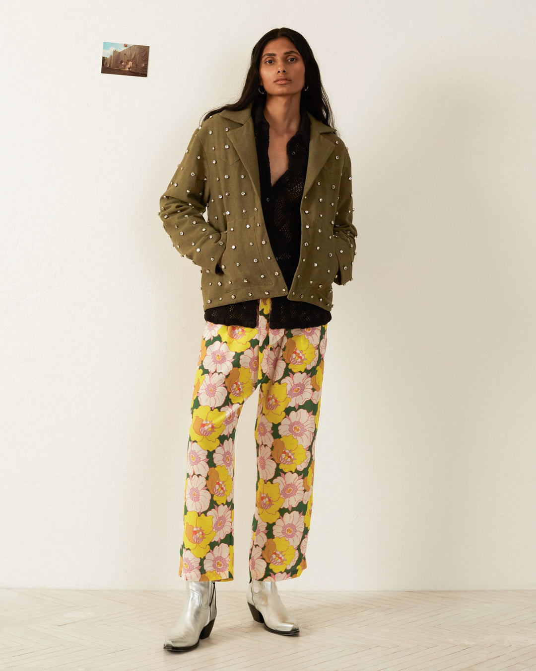 A person stands against a white wall, wearing the olive green Western Military Star Jacket with studs, a black top, and floral yellow and pink pants. They sport silver boots that catch the light like crystal-embellished gems. A small picture is pinned to the wall behind them.