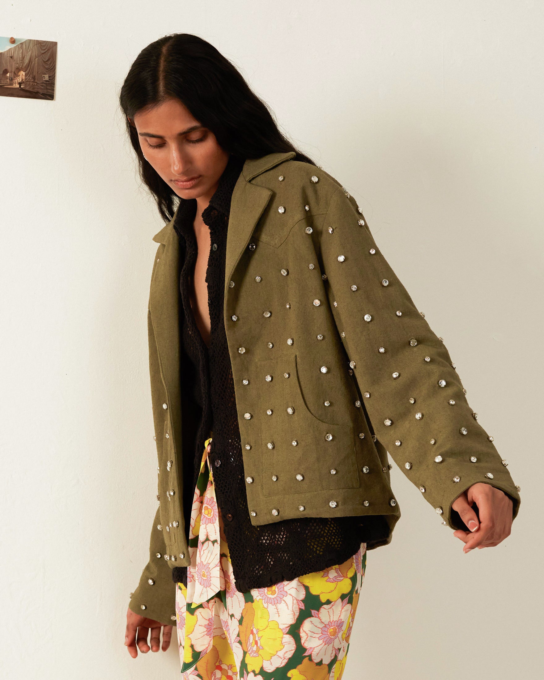 A person with long hair wears a Western Military Star Jacket in olive green silk, embellished with pearls, over a black top, and pairs it with floral yellow pants. They stand against a plain white wall with a small picture taped on it.