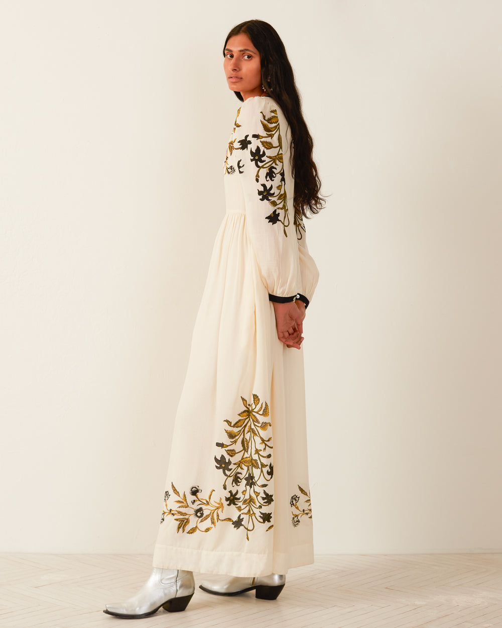 A person with long dark hair stands against a light background in the Winifred Olive Lily Valley Dress, showcasing its floral print and black and gold embroidery. They pair it with shiny silver boots and gaze to the side, exuding effortless elegance.